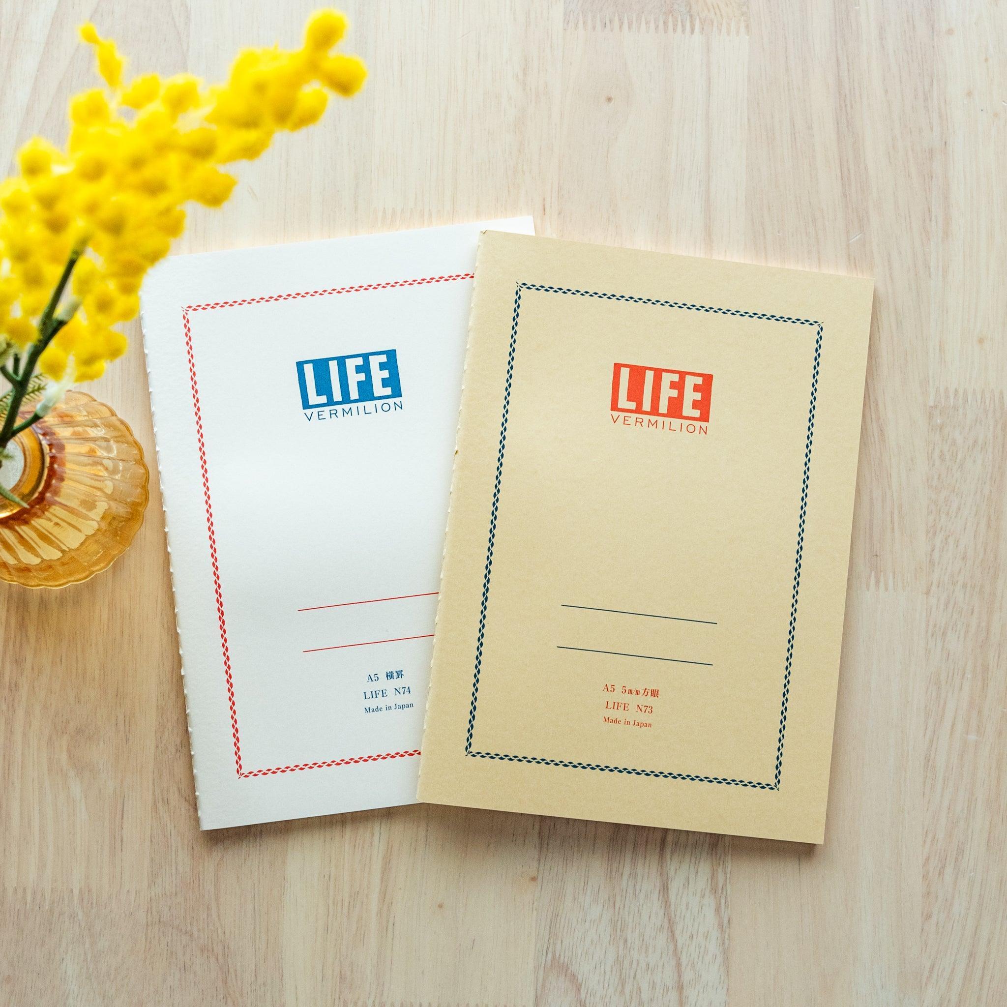 Two "LIFE Vermilion" notebooks in A5 size, one with a white cover and blue logo, the other beige with red logo, displayed on a wooden surface with yellow flowers.