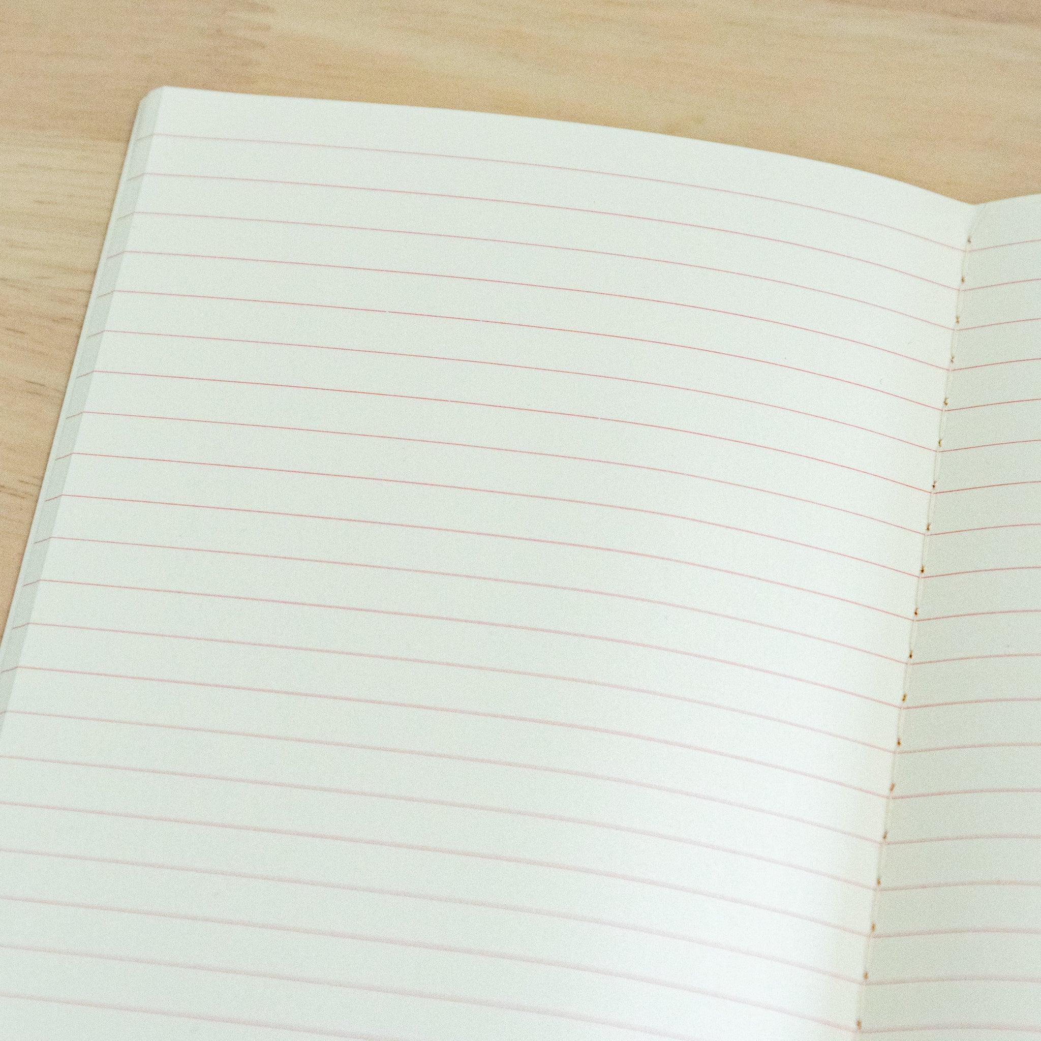 An open notebook with cream-colored lined paper featuring fine red line, displayed on a wooden surface.
