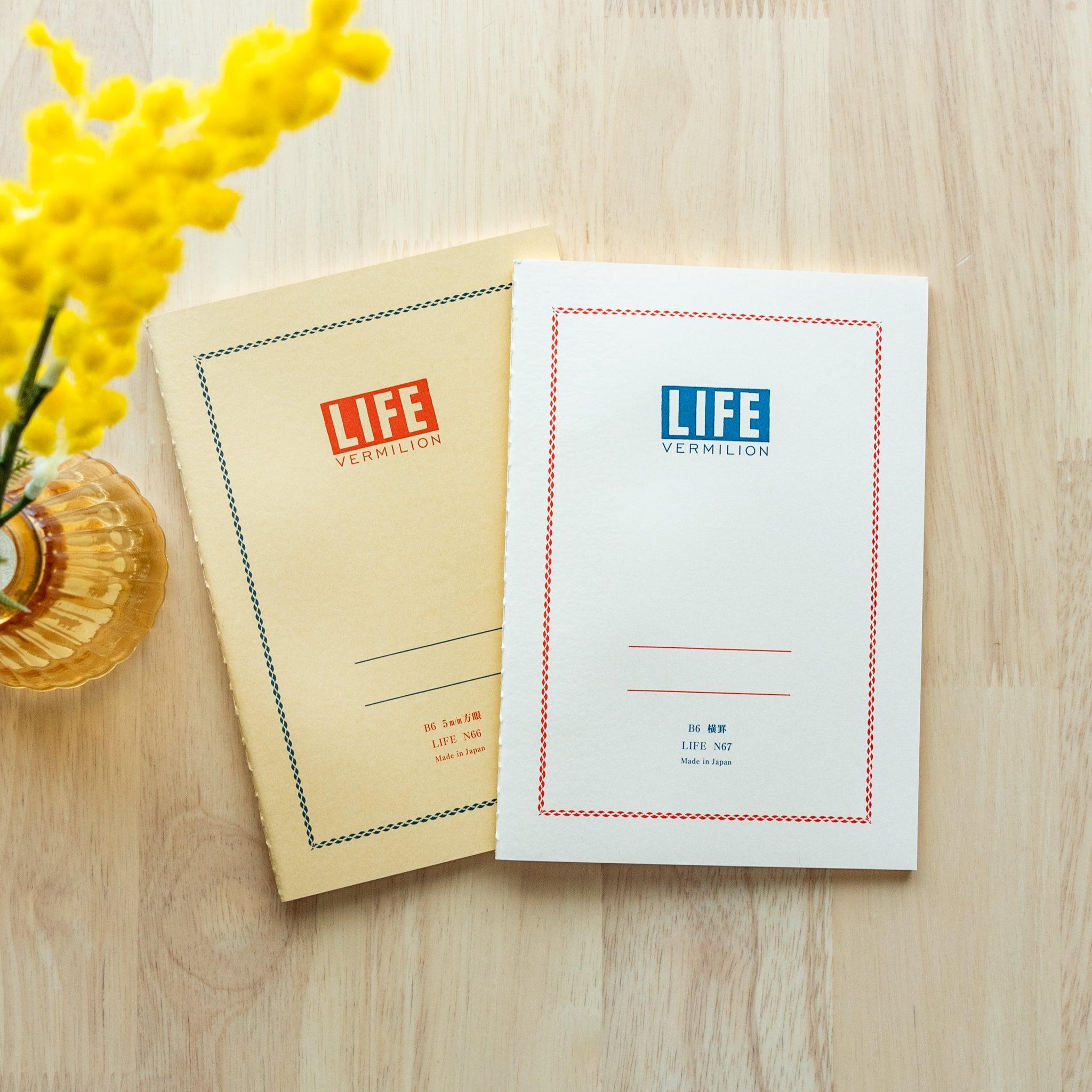 Two "LIFE Vermilion" notebooks in B6 size, one with a white cover and blue logo, the other beige with red logo, displayed on a wooden surface with yellow flowers.