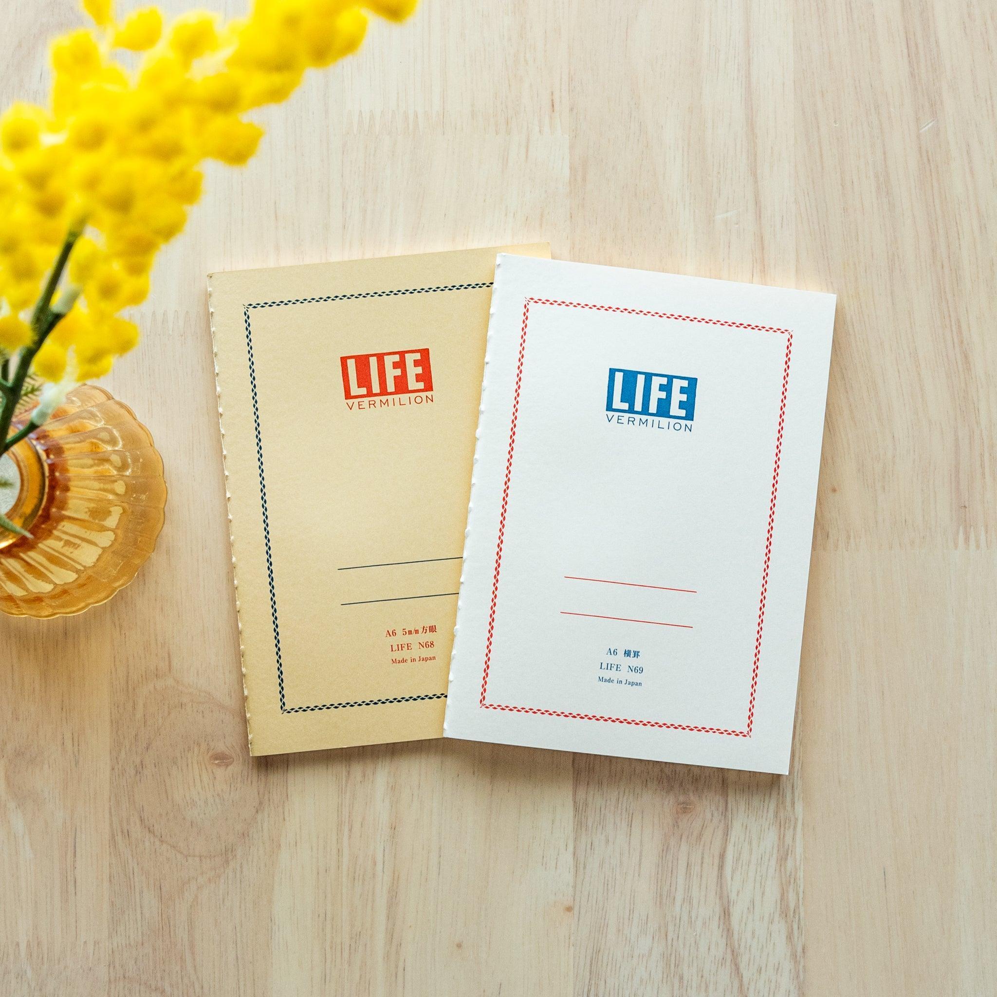 Two "LIFE Vermilion" notebooks in A6 size, one with a white cover and blue logo, the other beige with red logo, displayed on a wooden surface with yellow flowers.
