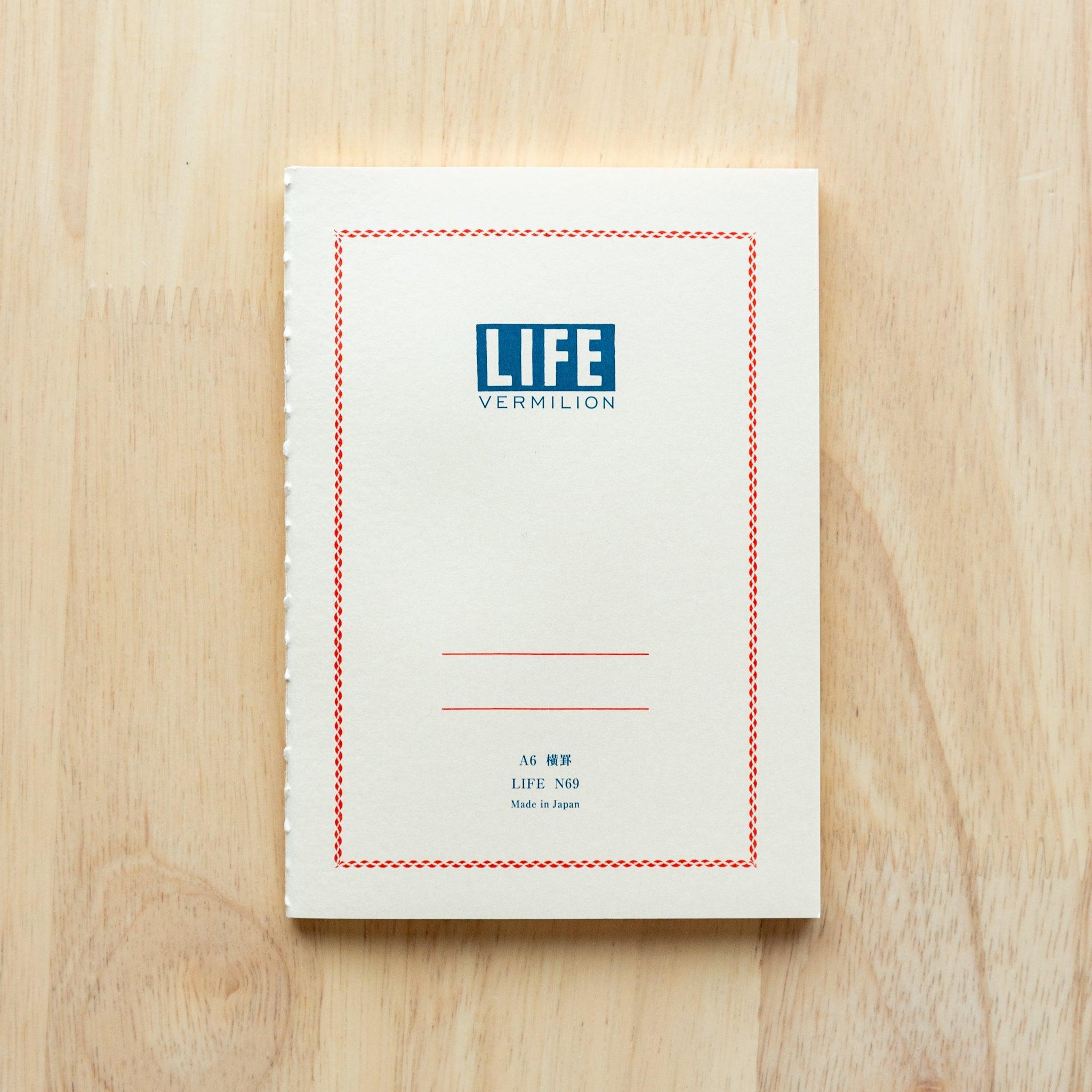 A single "LIFE Vermilion" notebook in A5, size with a white cover, blue logo, and minimal border design, displayed on a wooden surface.