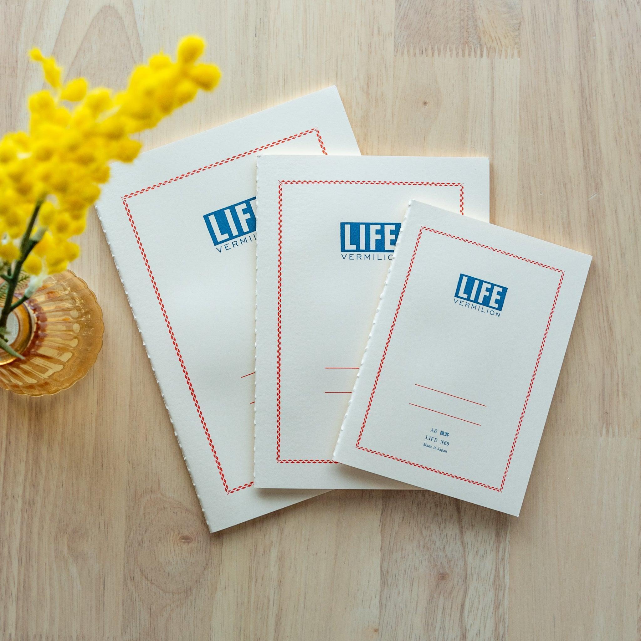 Three "LIFE Vermilion" notebooks in A5, B6 and A6 sizes, with white cover with blue logo, displayed on a wooden surface with yellow flowers.