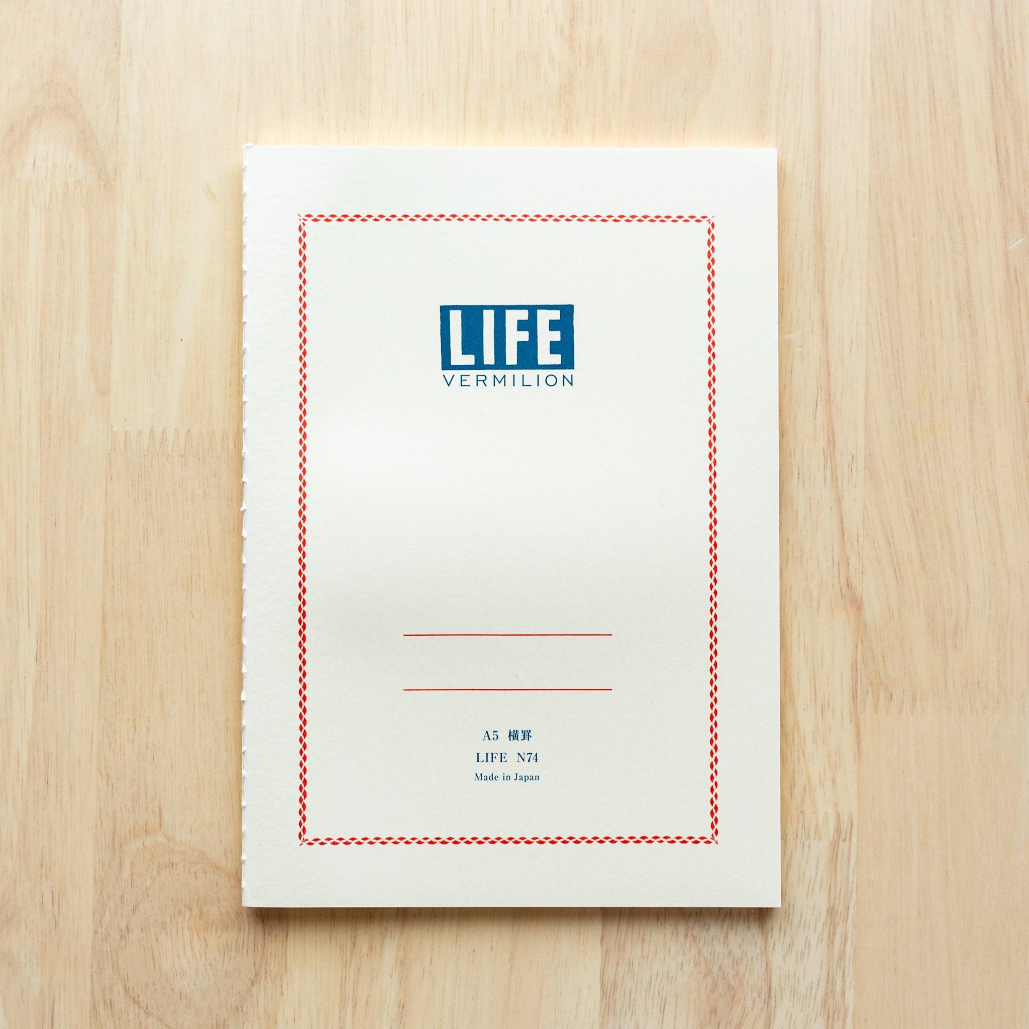 A single "LIFE Vermilion" notebook in A5 size with a white cover, blue logo, and minimal border design, displayed on a wooden surface.
