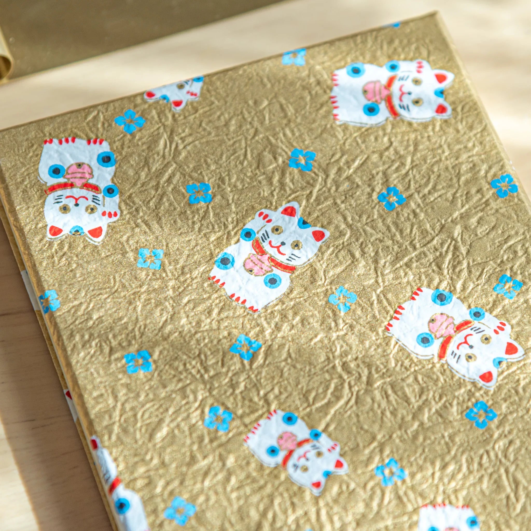 Gold textured washi cover close-up with white maneki-neko lucky cat figures and blue flowers, Japanese goshuincho stamp book