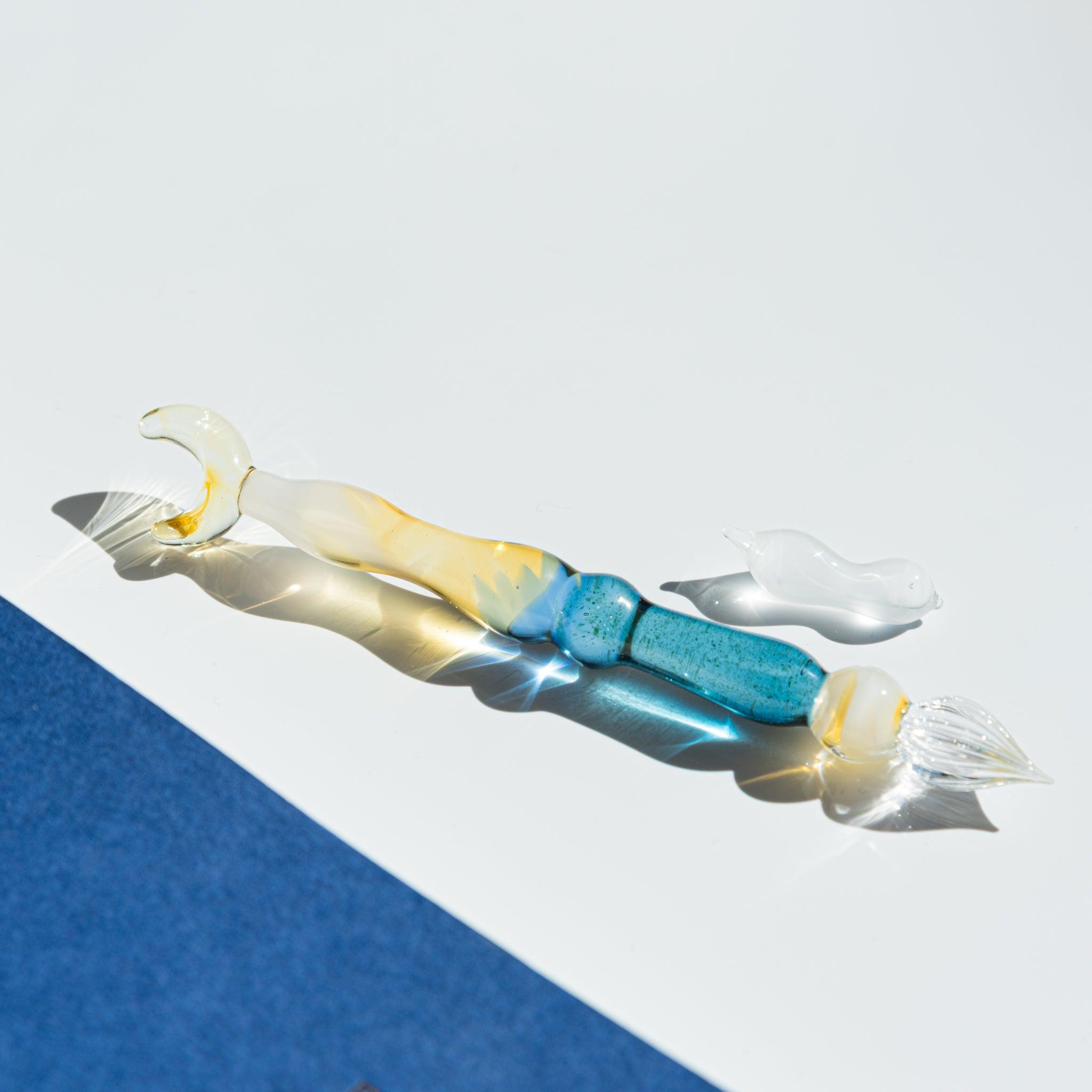 A glass pen with a crescent moon top and a blue gradient body lies on a reflective white surface. Its detachable nib rests beside it.
