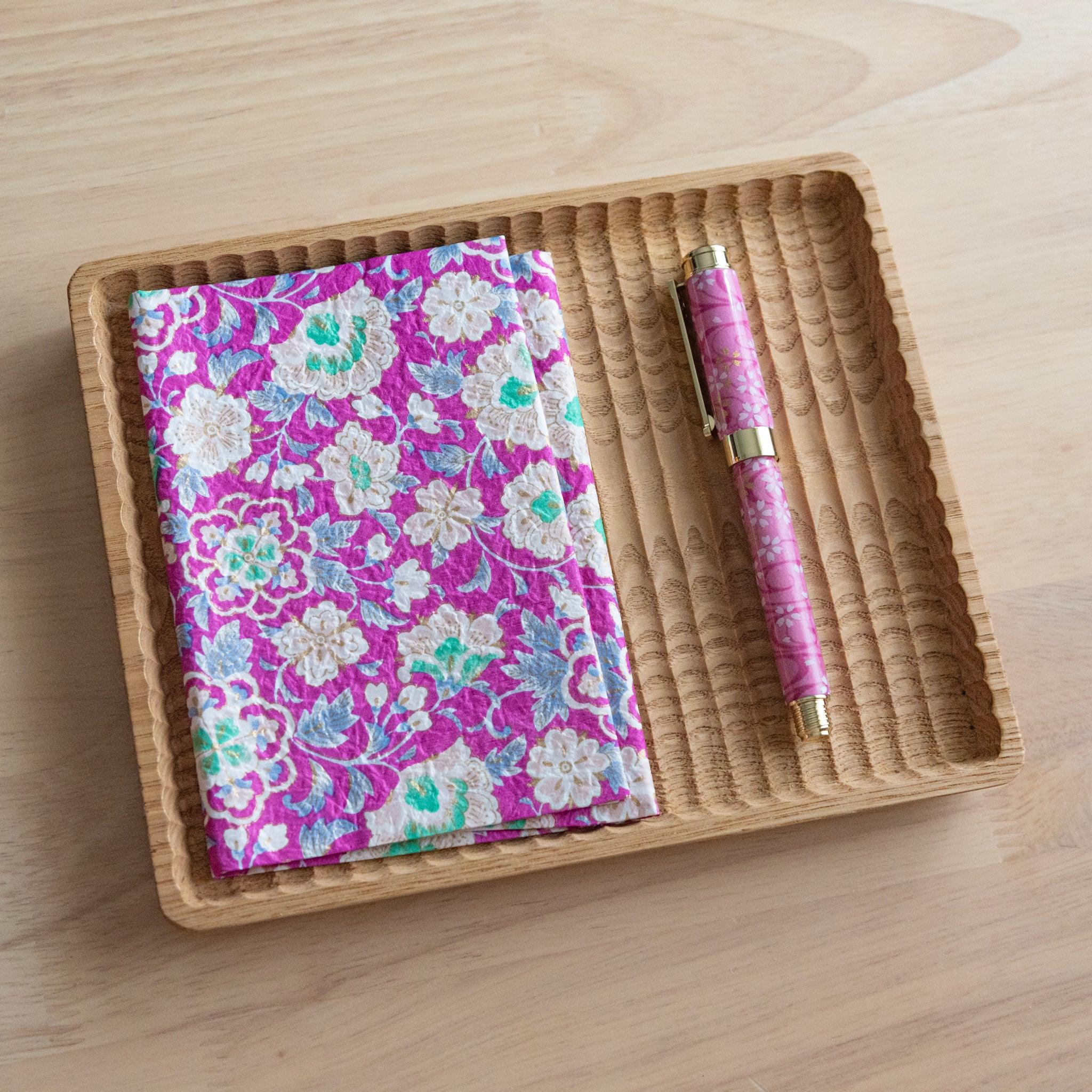 A notebook with a vibrant pink floral pattern and a matching pink pen, neatly arranged on a wooden tray on a light wooden surface.