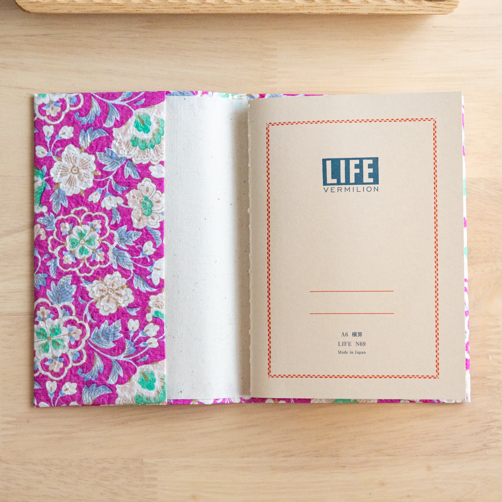 An open notebook with a vibrant pink floral cover, showing its title page labeled "Life Vermilion," placed on a wooden surface.