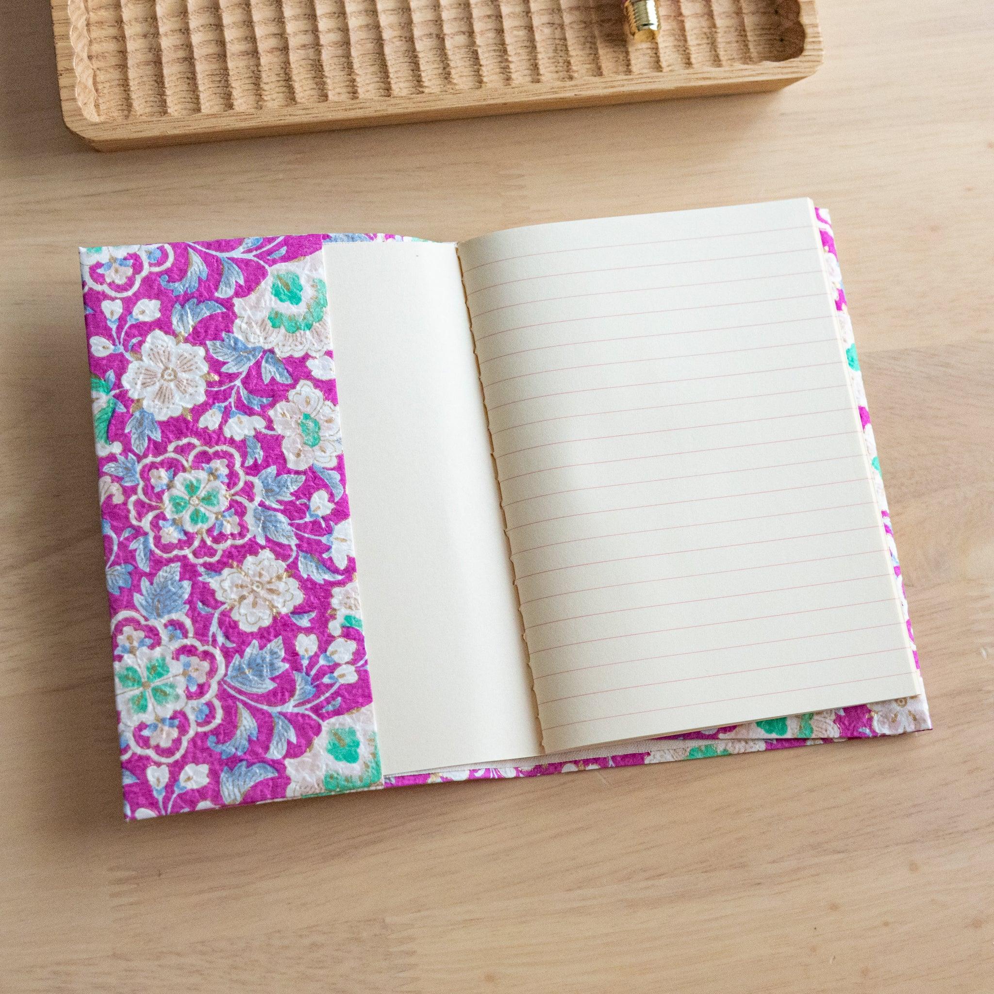 An open notebook with a vibrant pink floral cover, revealing one lined page and one blank page, placed on a light wooden surface.