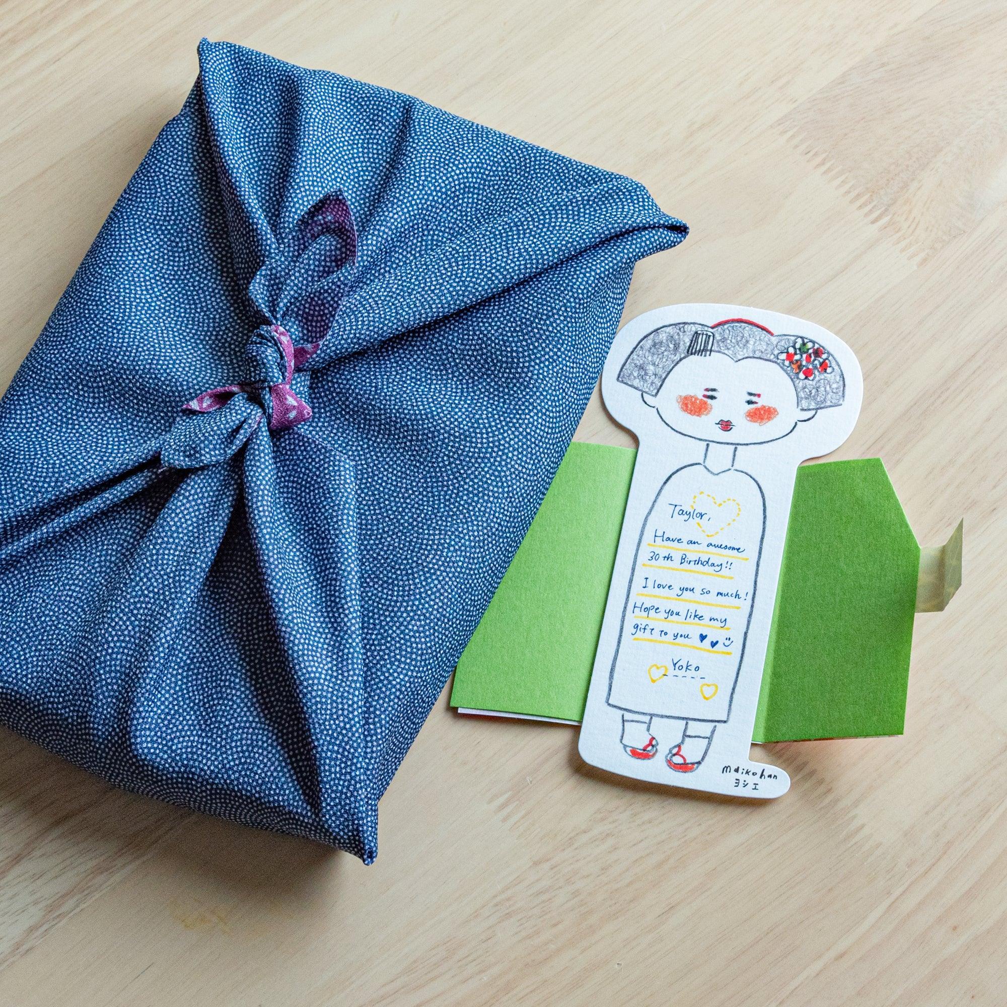 Open maiko die-cut message card with handwritten birthday note, beside a furoshiki-wrapped gift and green envelope on wood