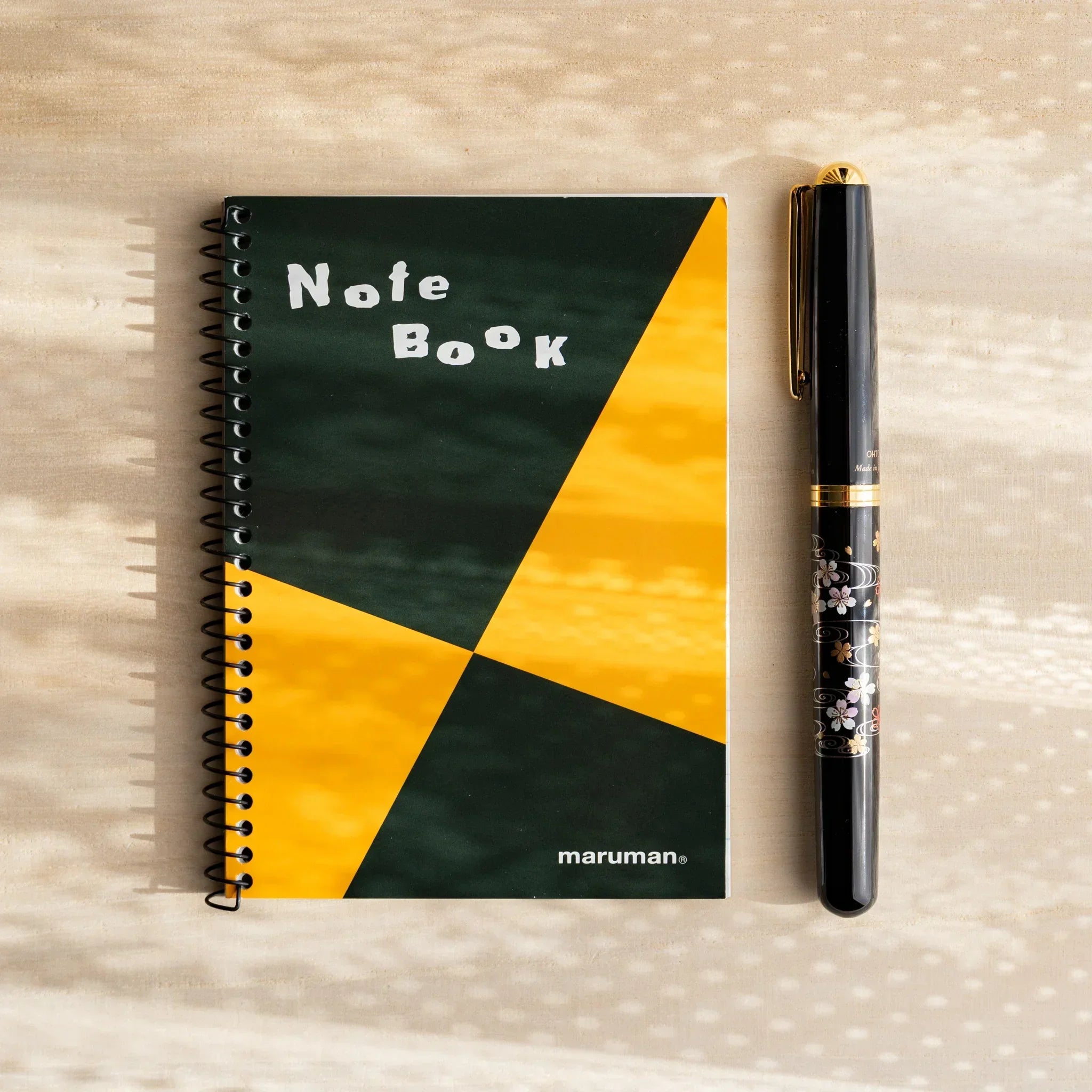 Japanese notebook by maruman with orange and green green spiral-bound cover, paired with a black pen featuring floral motifs.