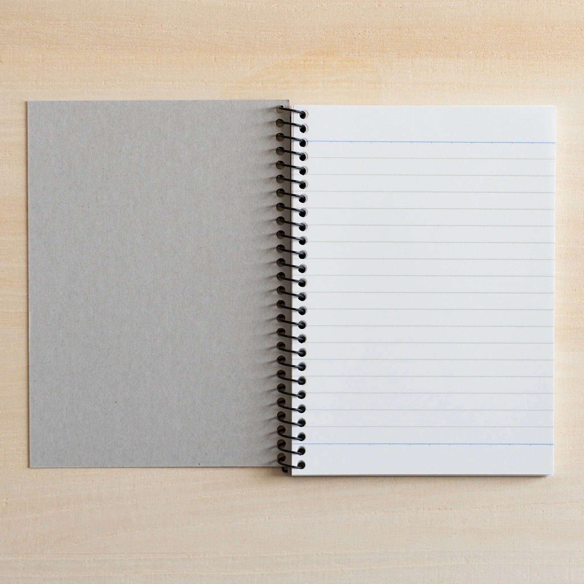 Japanese notebook by maruman with spiral binding, open to lined white pages with a grey cover, resting on a light wood desk.