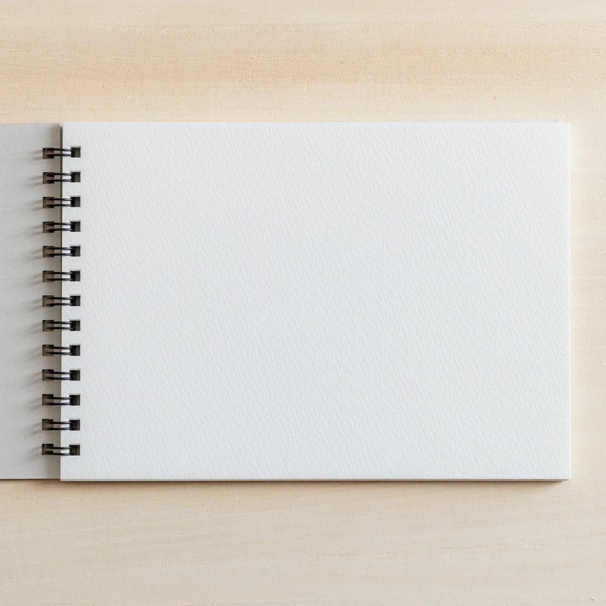 A maruman Japanese sketchbook with blank white cover and ring binding on the left sits on a light wooden desk for arts and crafts.