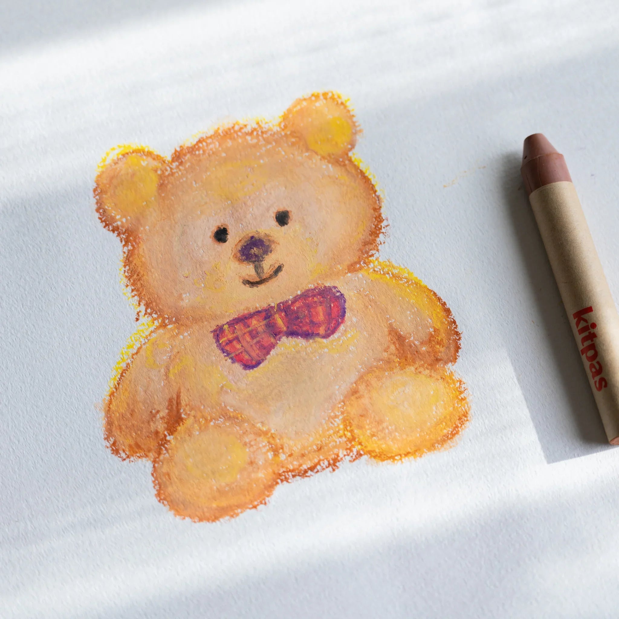 A maruman Japanese sketchbook page with a crayon drawing of a teddy bear with a red bow tie and a beige crayon beside it.