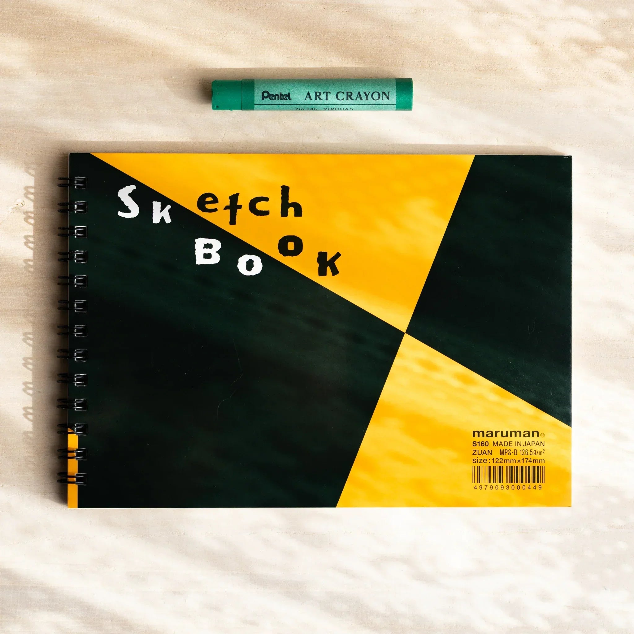A maruman Japanese sketchbook with yellow and black diagonal cover and ring binding rests on a desk with a green crayon.