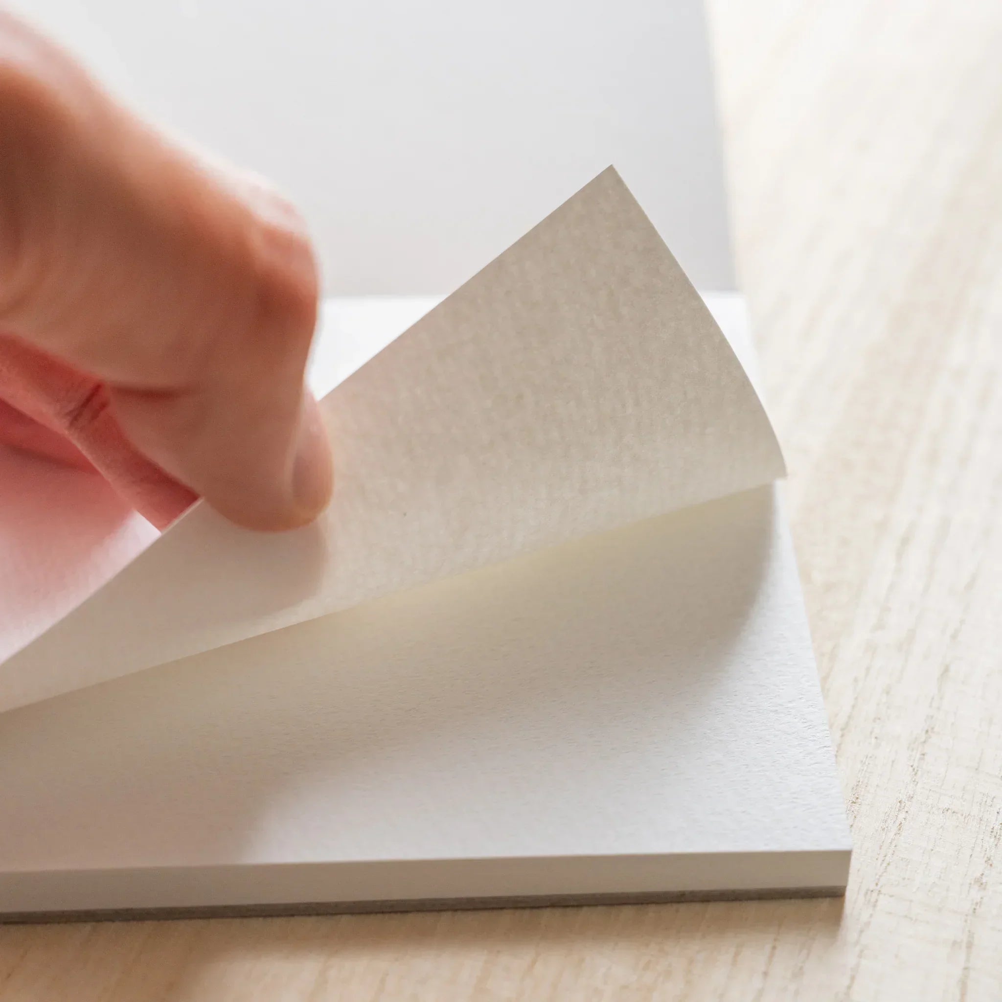 Hand lifting a sheet in a Maruman Japanese sketchbook to show thick textured white paper on a wooden surface.