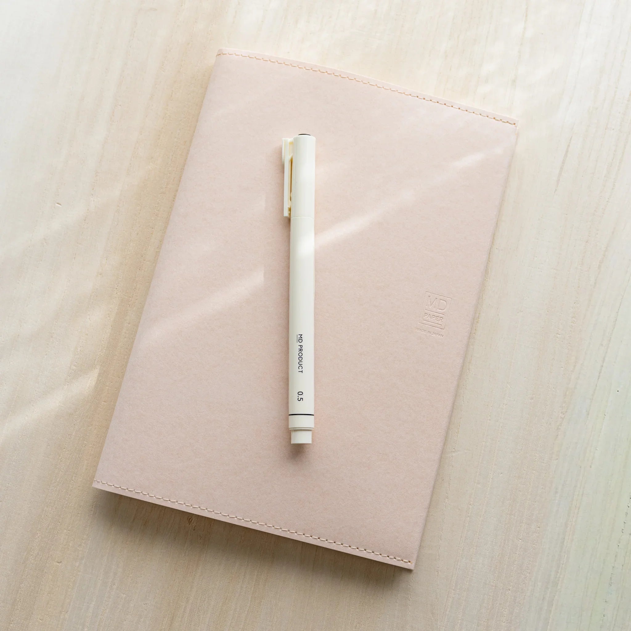 Midori MD Notebook Paper Cover with notebook inside on wood, white MD pen placed diagonally on pale pink paper cover