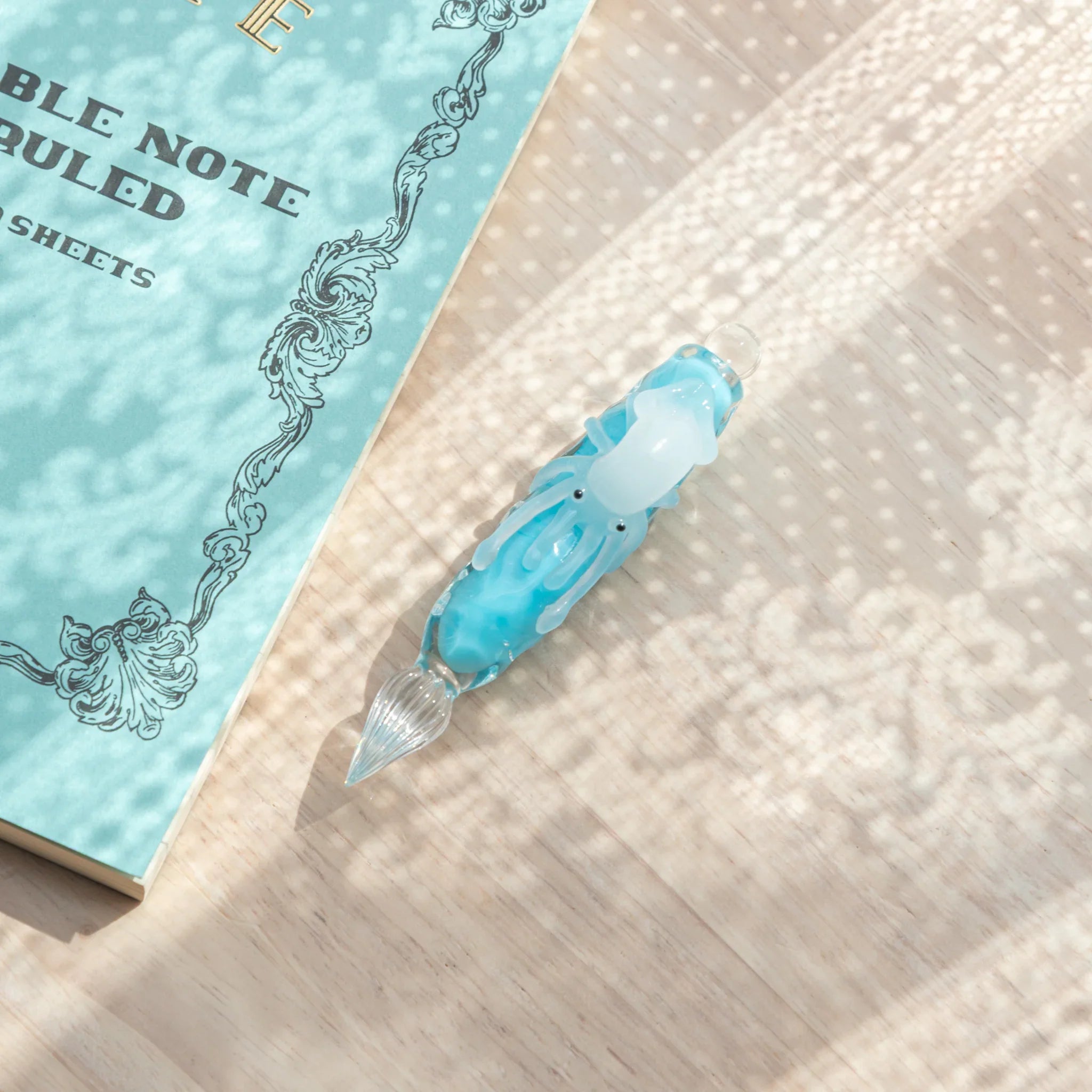 Overhead view of light blue glass dip pen on wood surface showing white squid and glass nib, LIFE Noble Note notebook beside