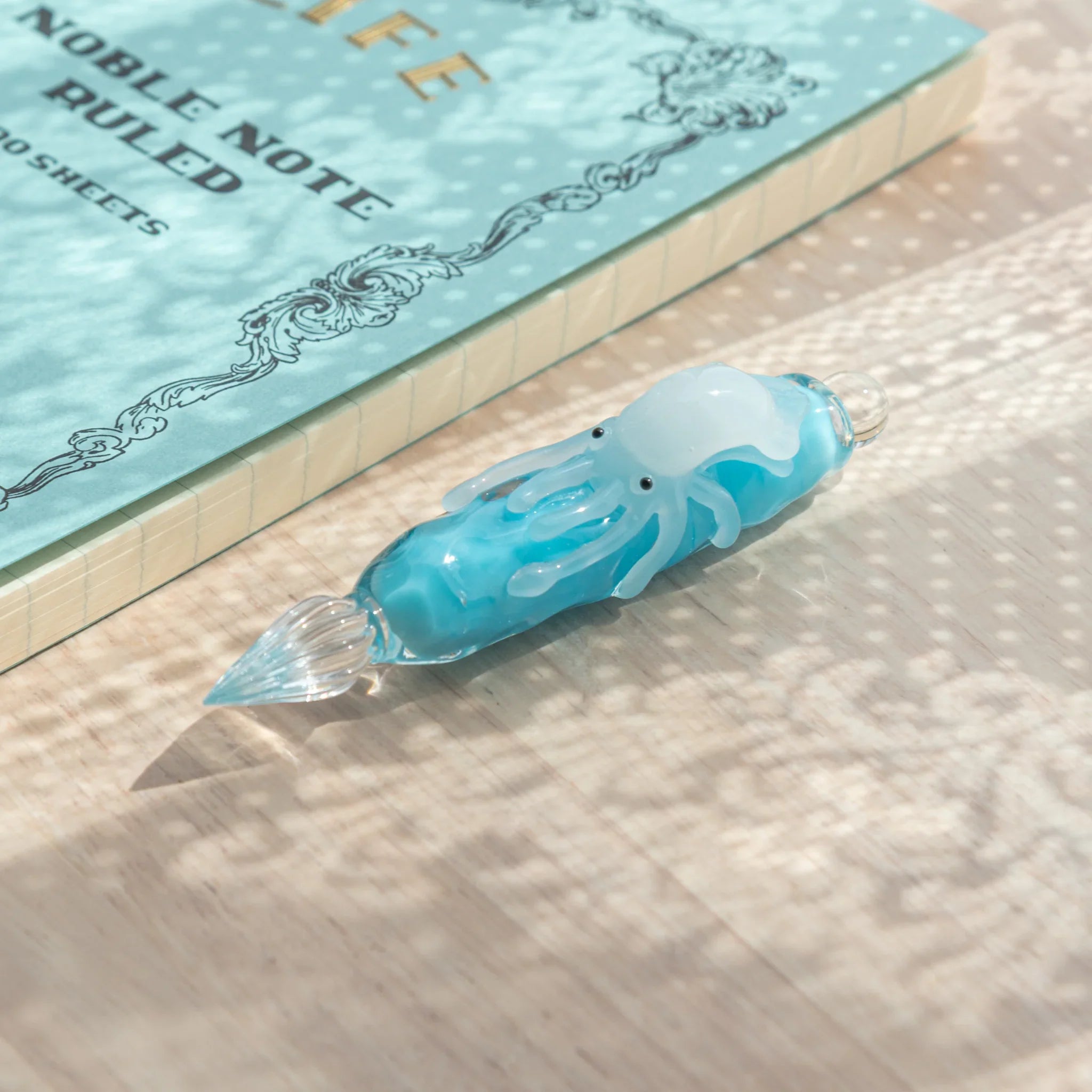 Light blue glass dip pen with white squid figure lying on wood surface, glass nib at left, LIFE Noble Note notebook behind