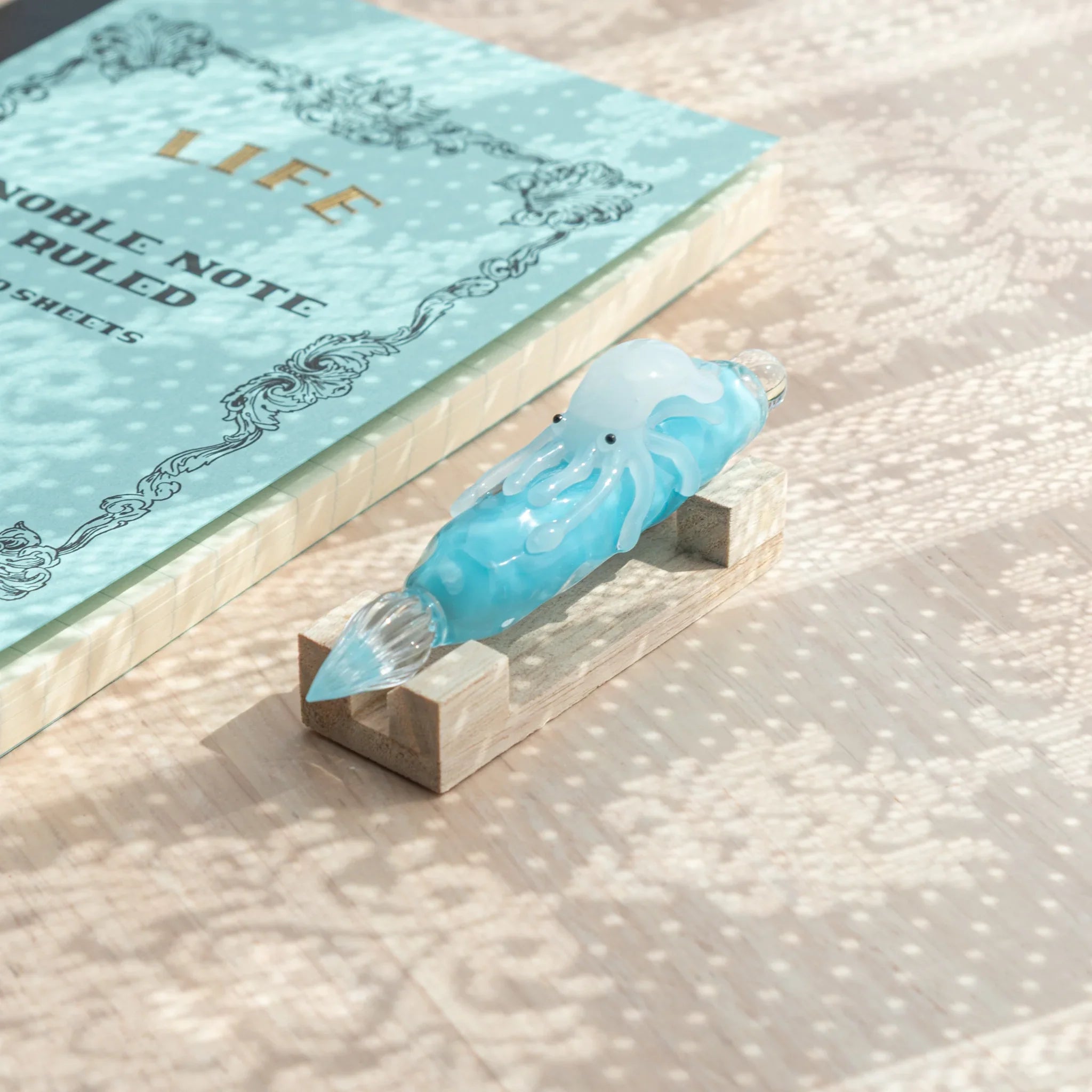 Light blue glass dip pen with white squid figure on natural wood pen stand, LIFE Noble Note Ruled notebook leaning behind