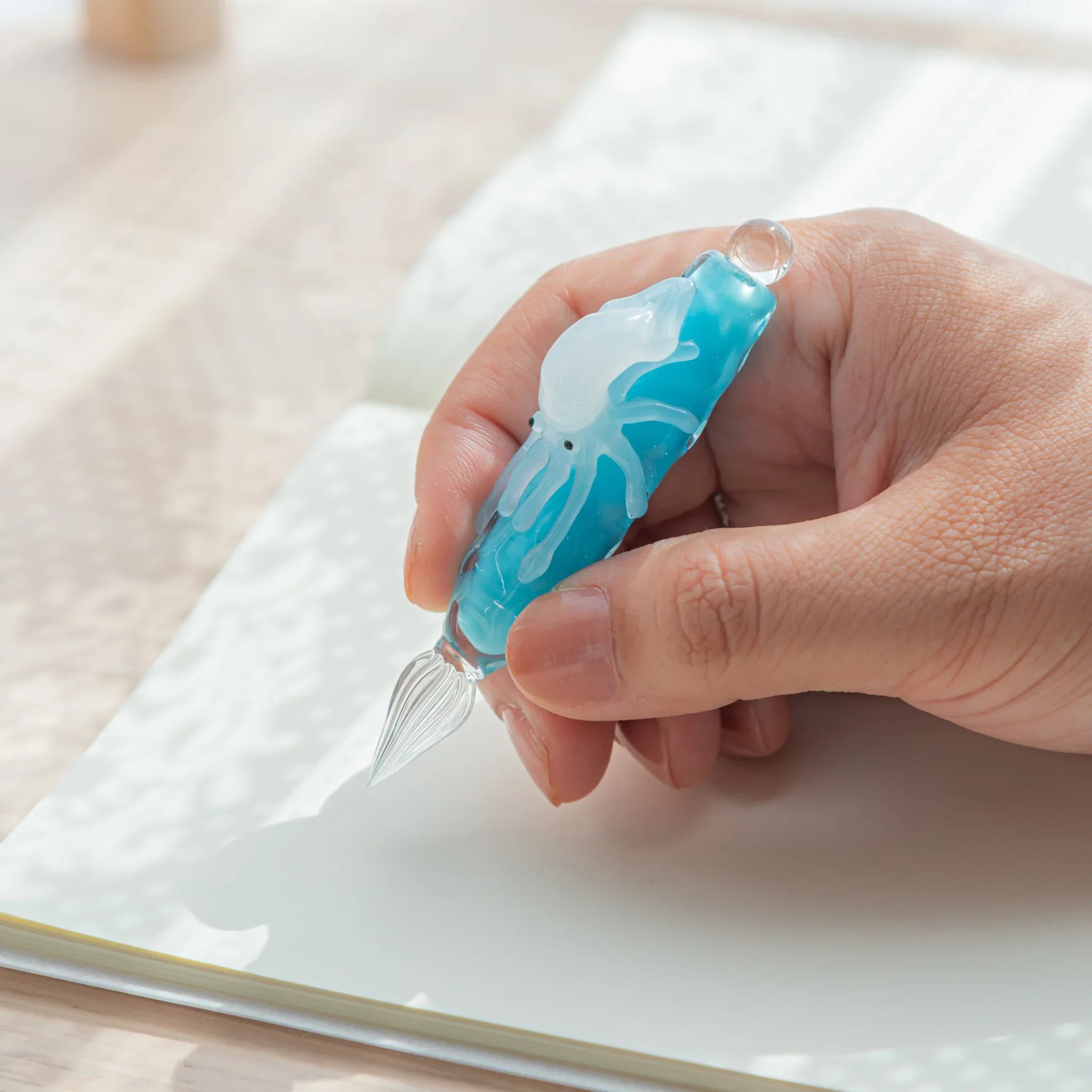 Hand holding light blue glass dip pen over open notebook, showing white squid figure and ribbed glass nib