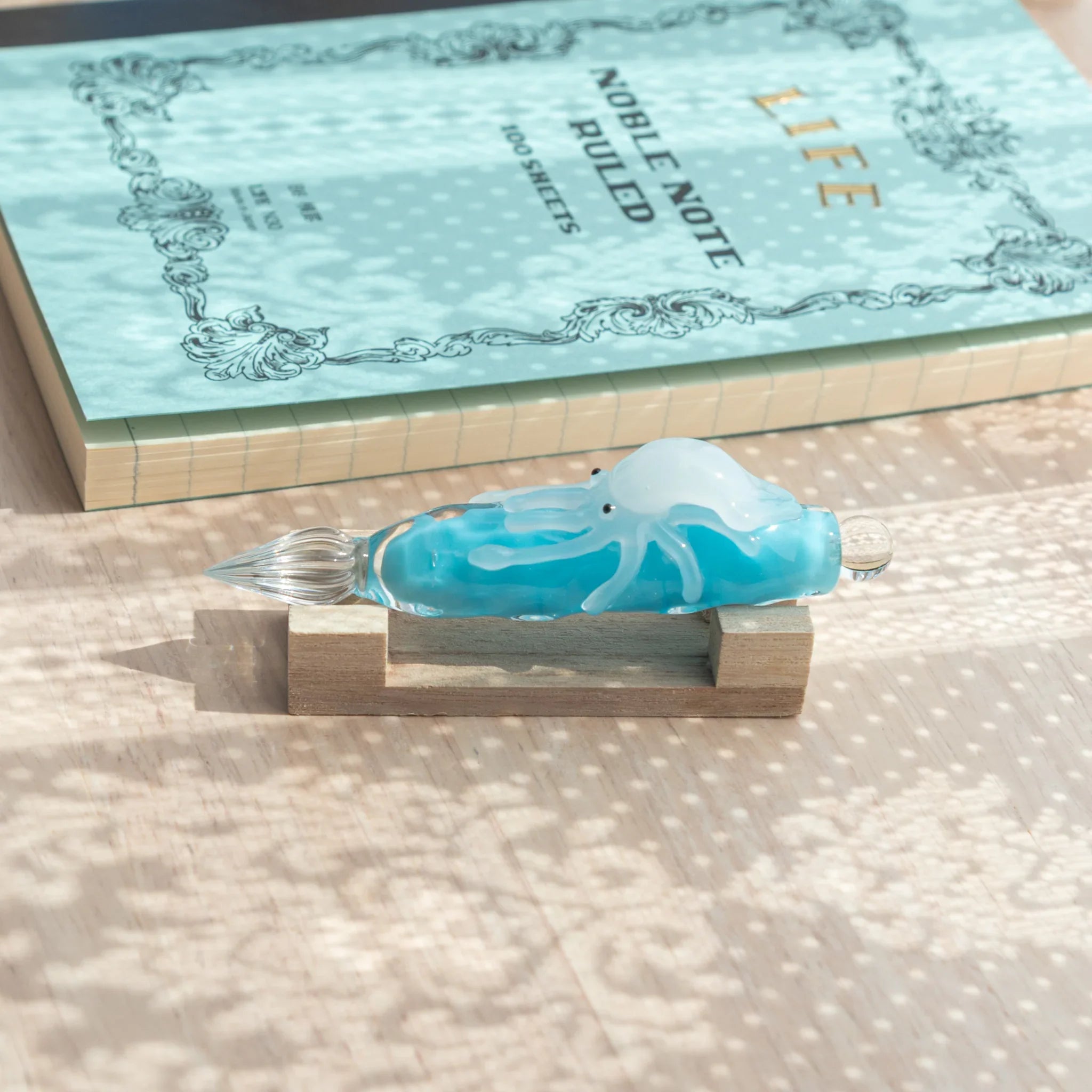 Light blue glass dip pen on wood pen stand beside flat LIFE Noble Note Ruled notebook, showing white squid figure