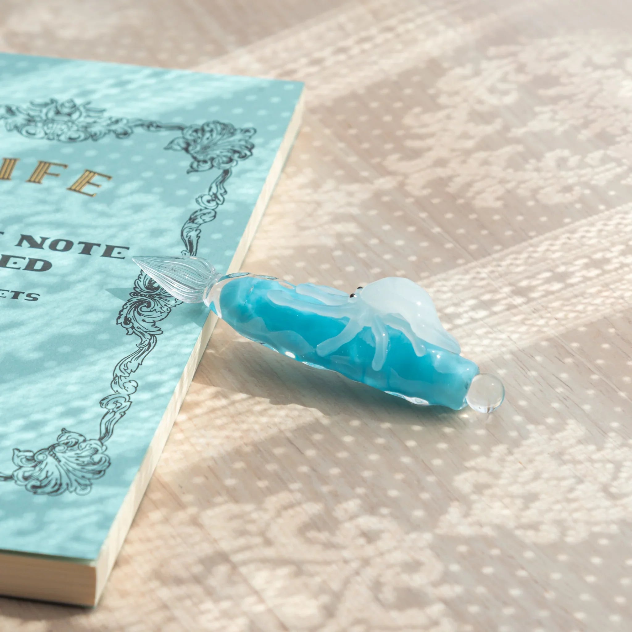 Light blue glass dip pen on wood surface beside LIFE Noble Note notebook, showing white squid figure and glass nib