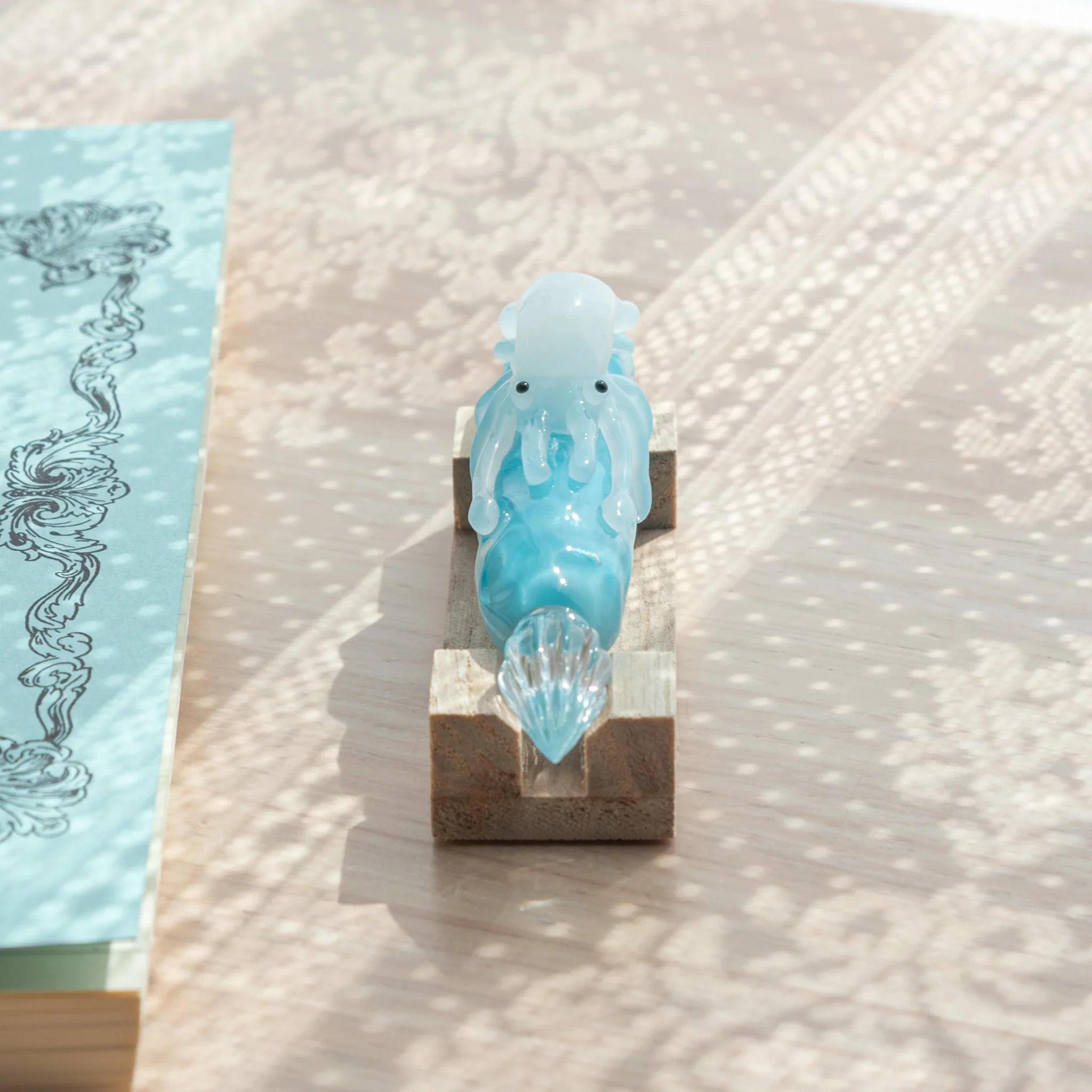 Front-end view of light blue glass dip pen on wood pen stand showing clear glass nib and white squid figure facing forward