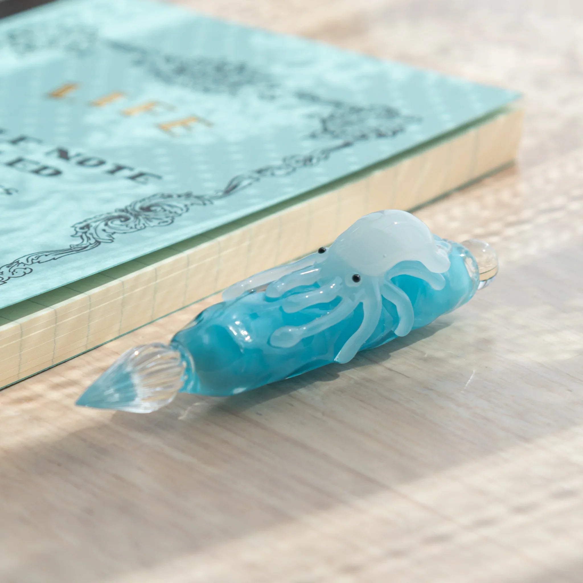 Light blue glass dip pen on wood surface showing white squid figure and glass nib, LIFE Noble Note notebook behind