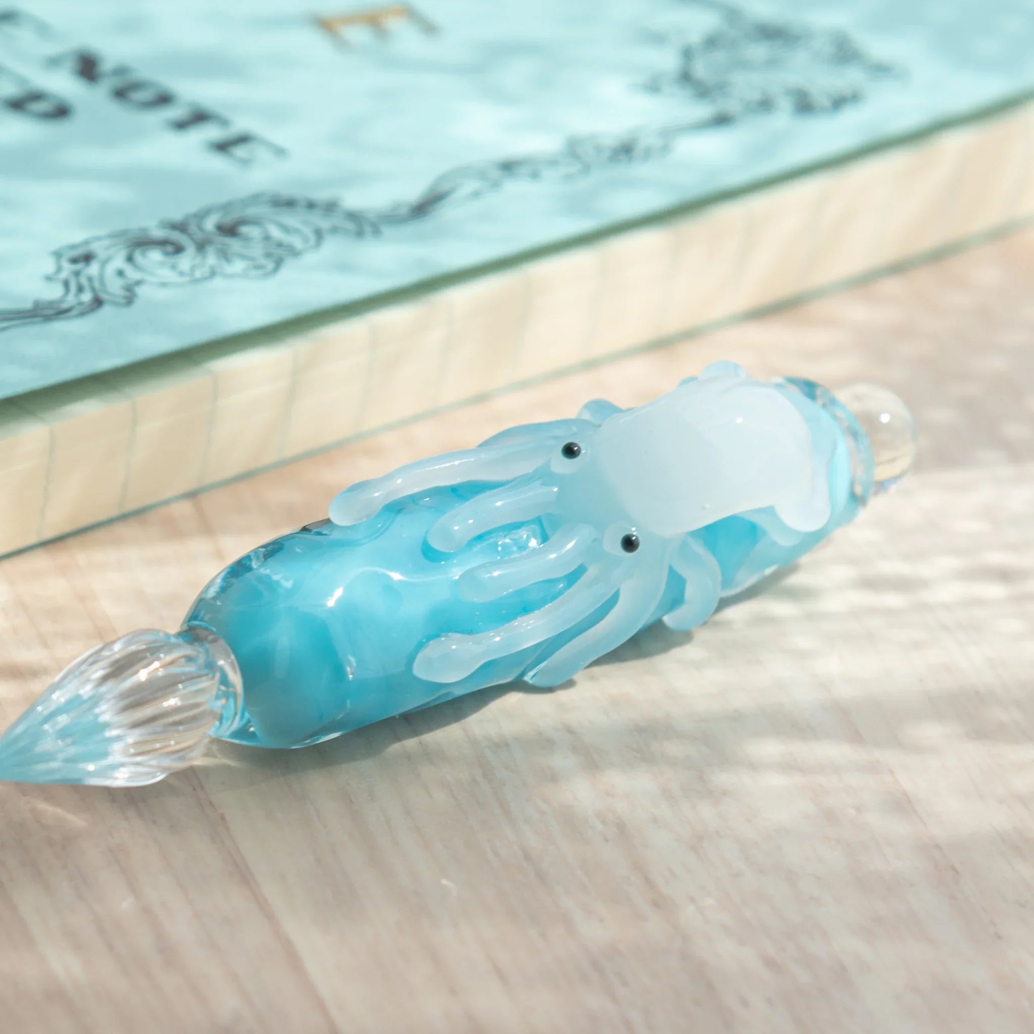Close-up view of light blue glass dip pen with white squid showing black eyes and tentacles on barrel, glass nib below