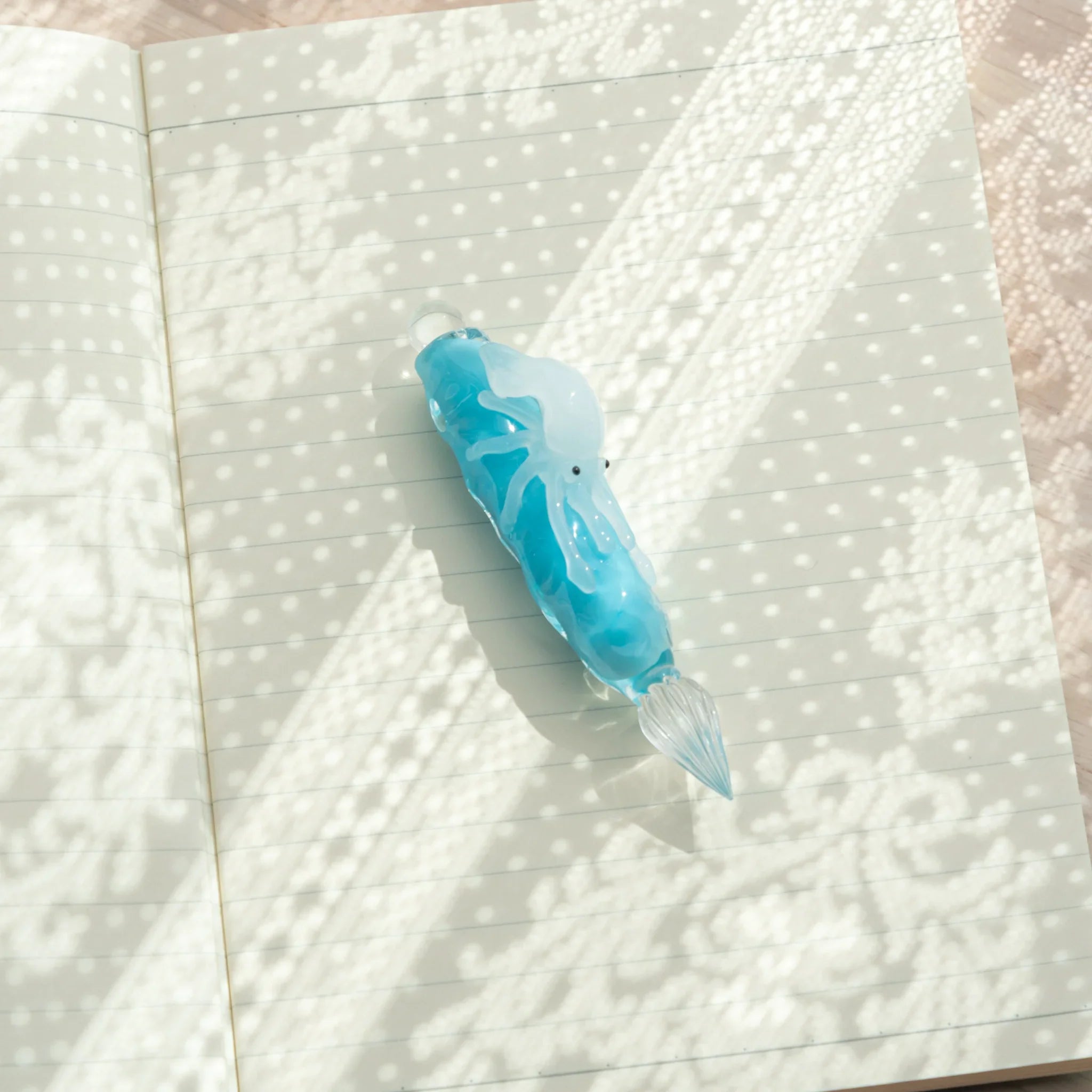 Light blue glass dip pen with white squid figure lying on open ruled notebook pages, viewed from above