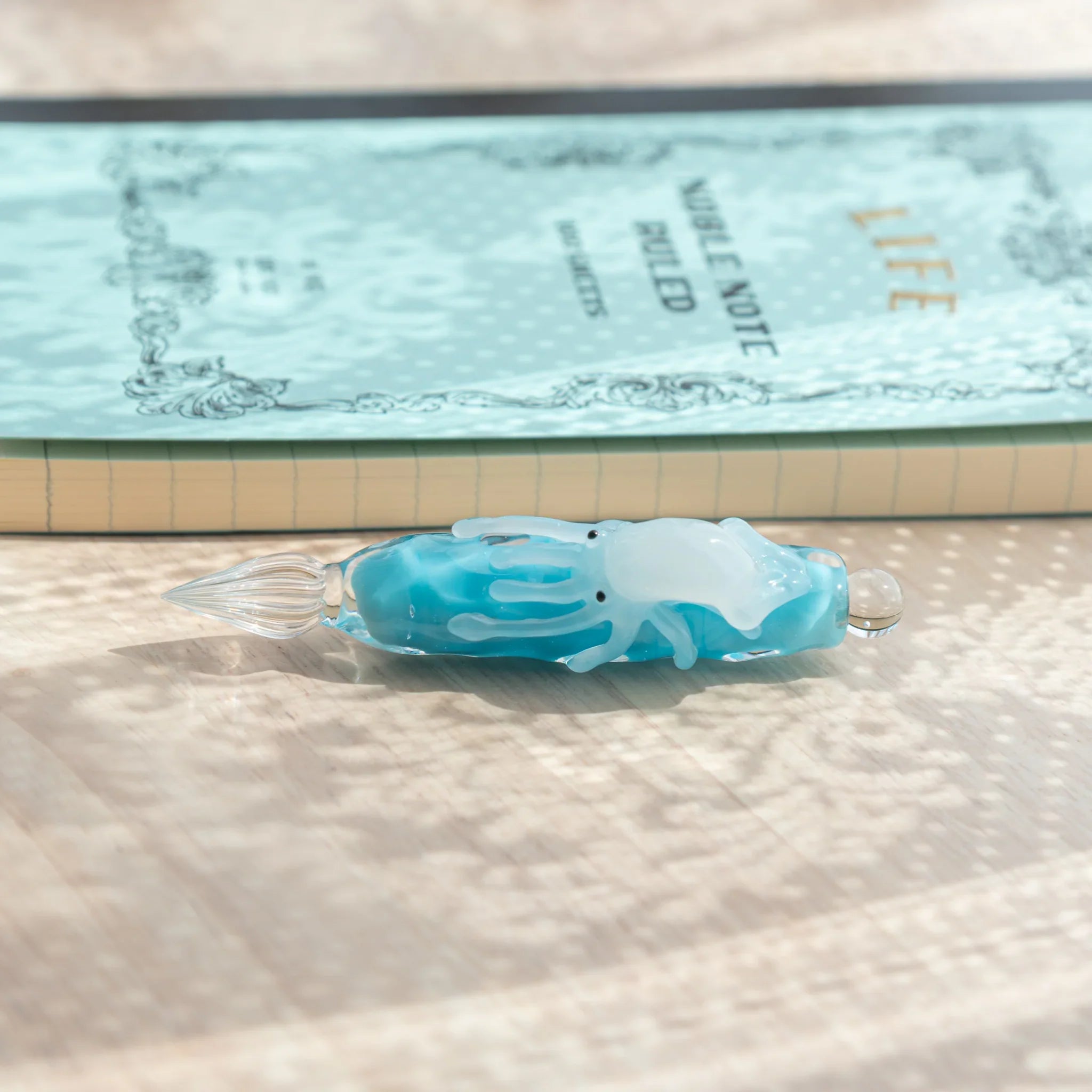 Top-down view of light blue glass dip pen showing white squid figure and glass nib, LIFE Noble Note notebook behind