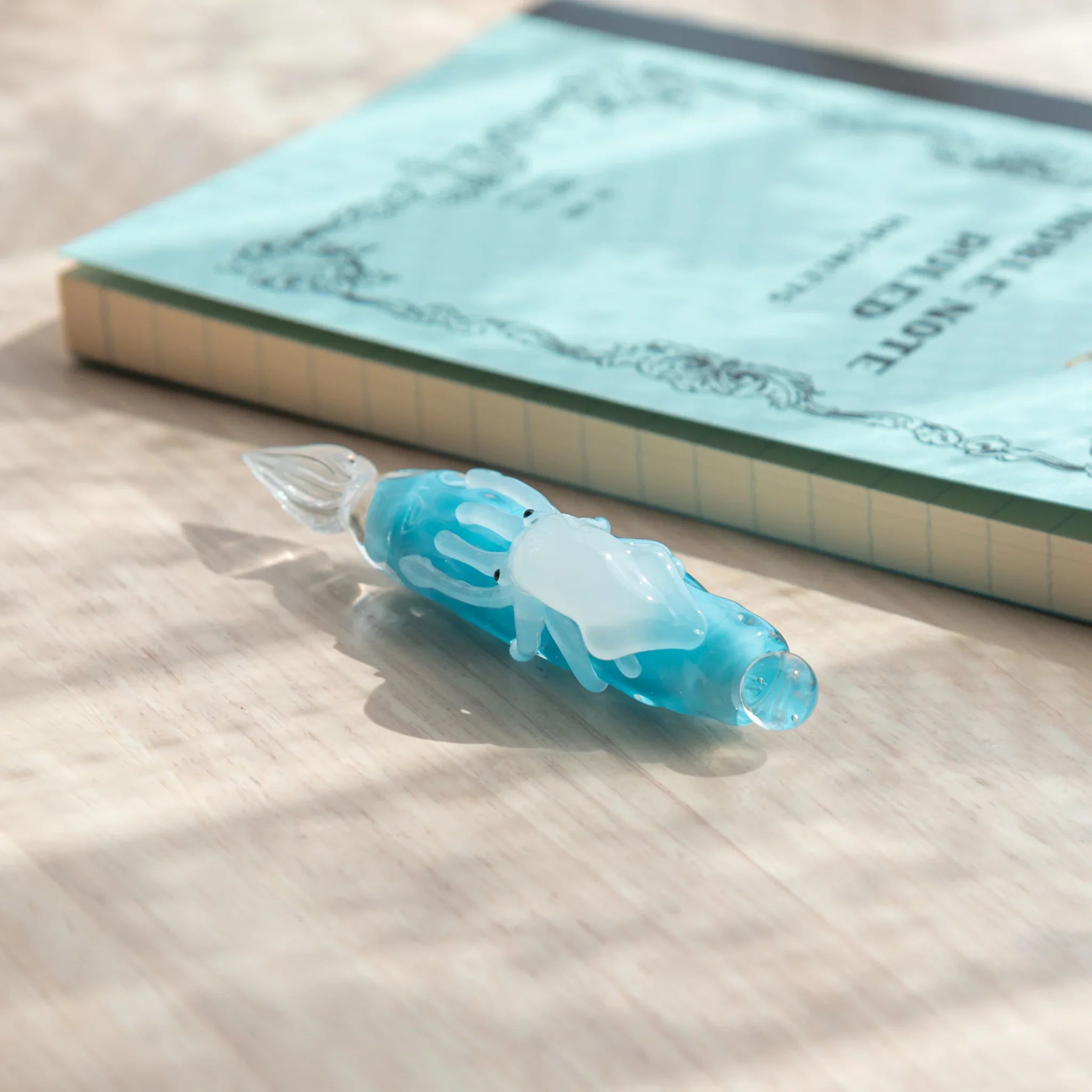 Light blue glass dip pen beside upright LIFE Noble Note Ruled notebook, showing white squid figure on barrel and glass nib