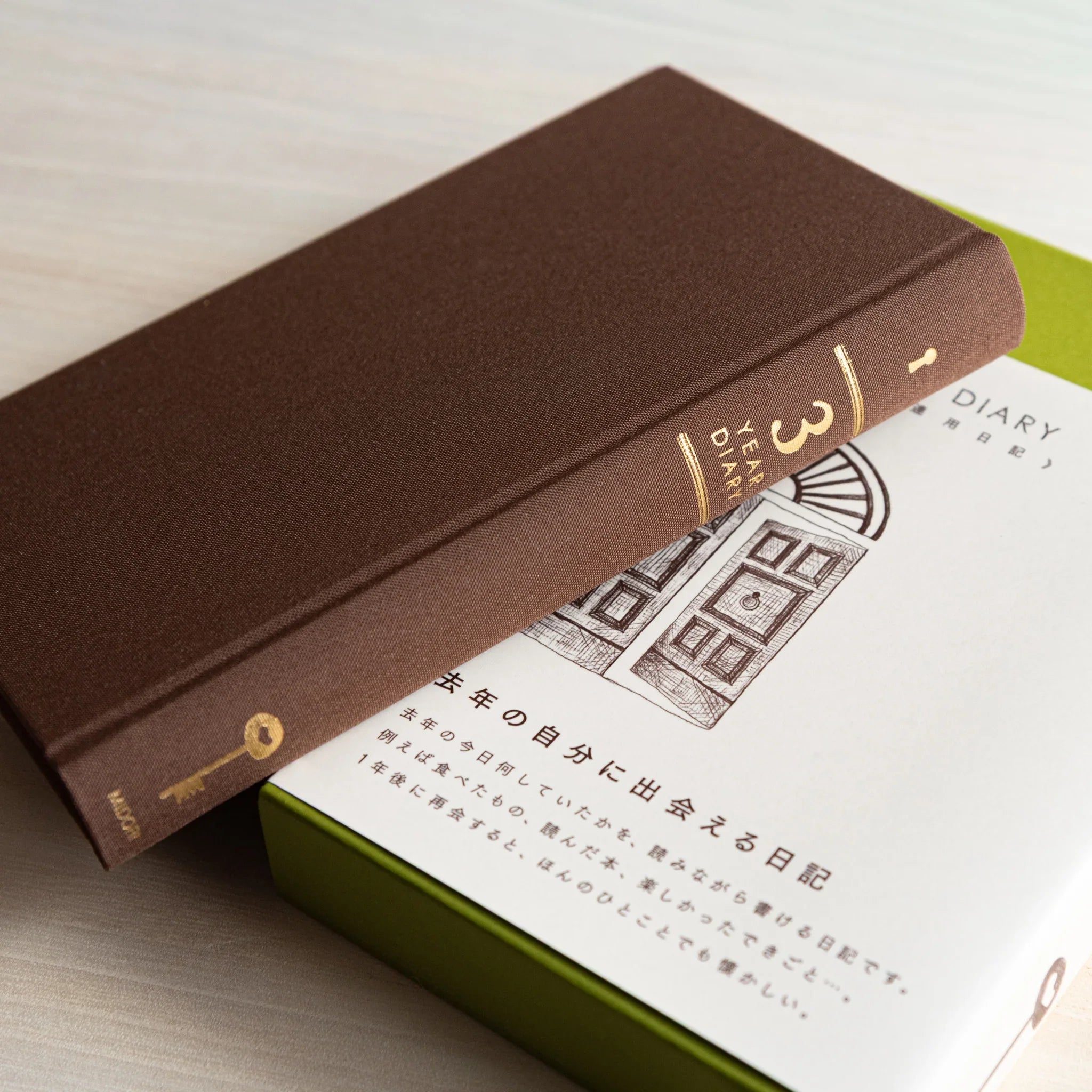 Angled view of brown Midori Japanese journal “3 Year Diary Gate Design” resting on a white and green slipcase.
