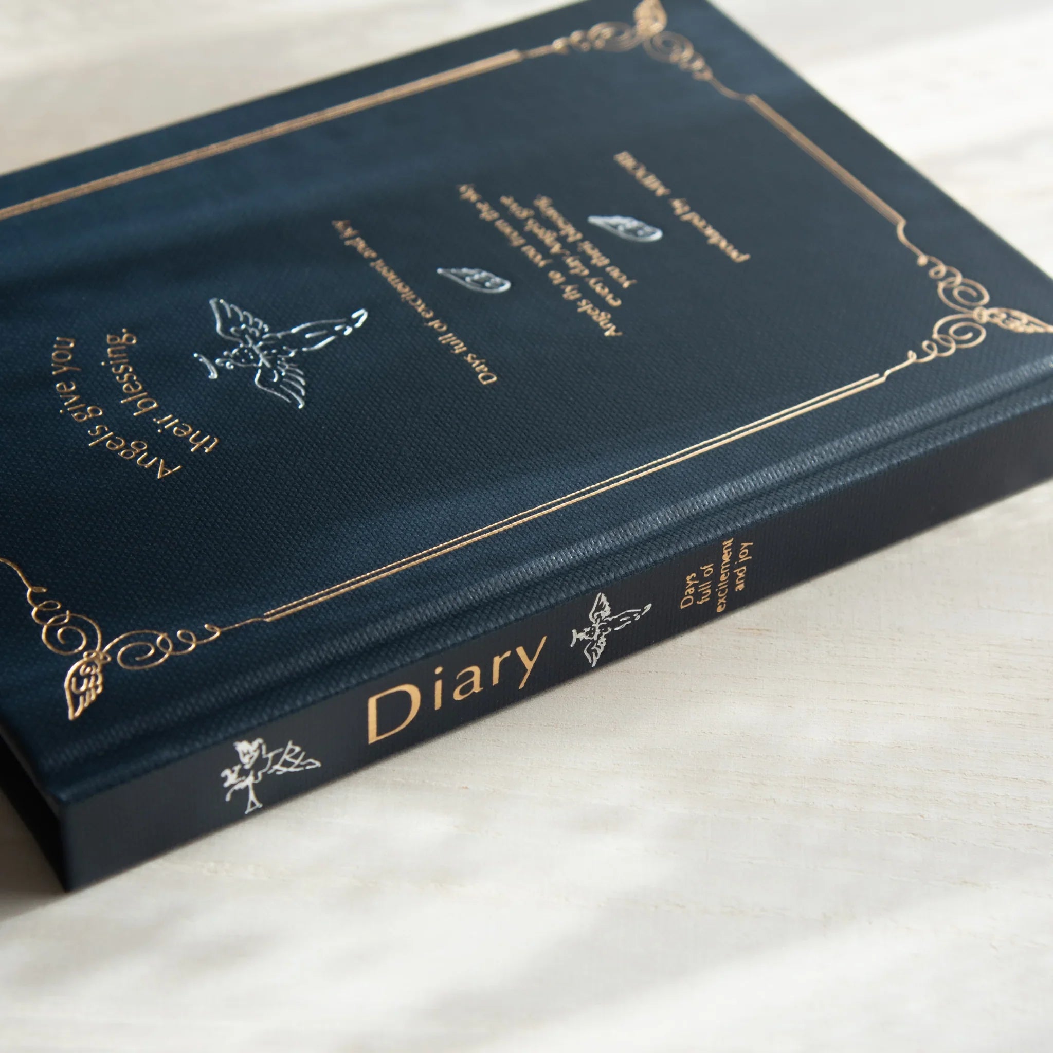Midori Angel Motif Journal on cream surface, showing spine labeled 'Diary', navy cover with gold foil angel design and quote