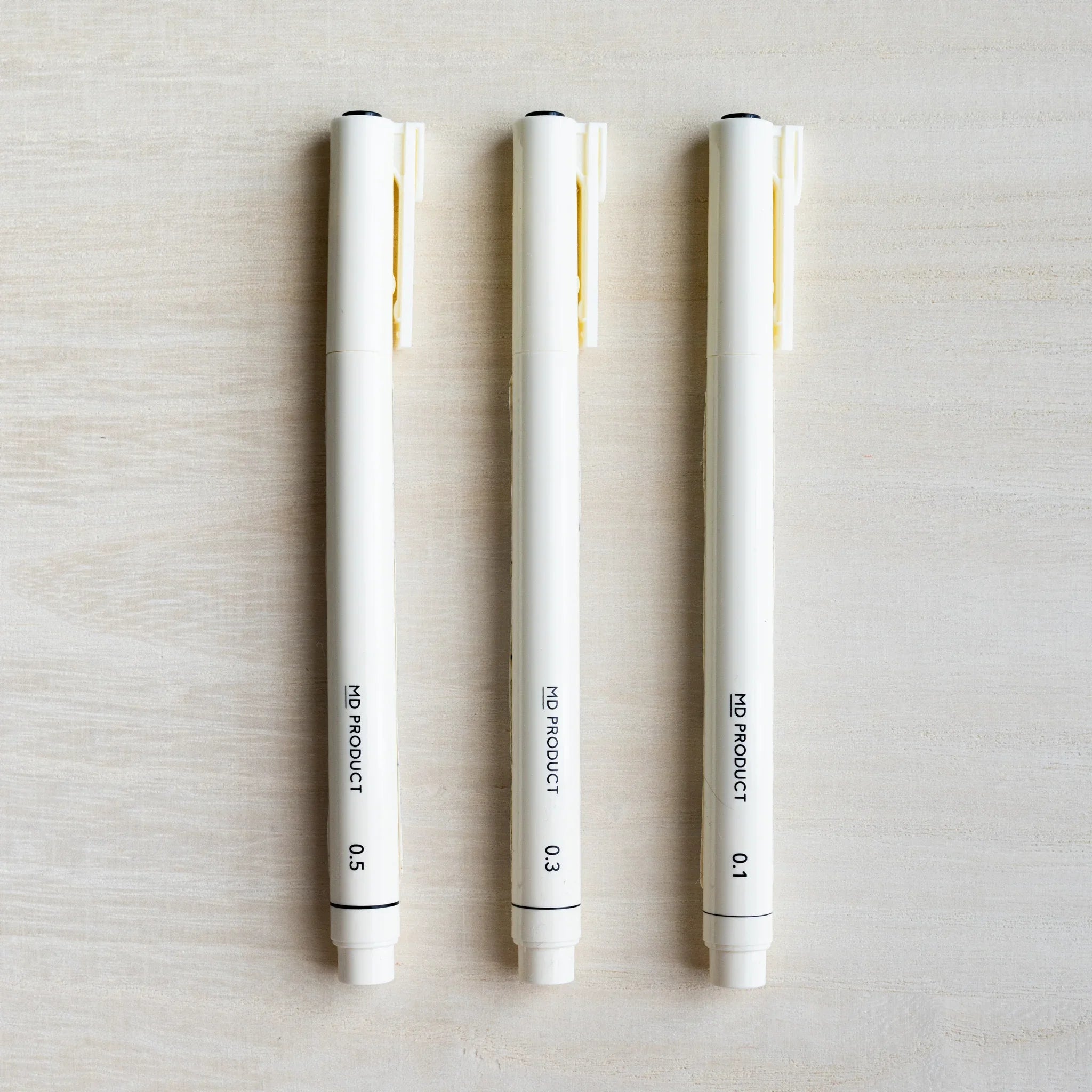Three Midori MD Drawing Pens capped, ivory barrels marked 0.5, 0.3, and 0.1 mm, clips visible, on pale wood