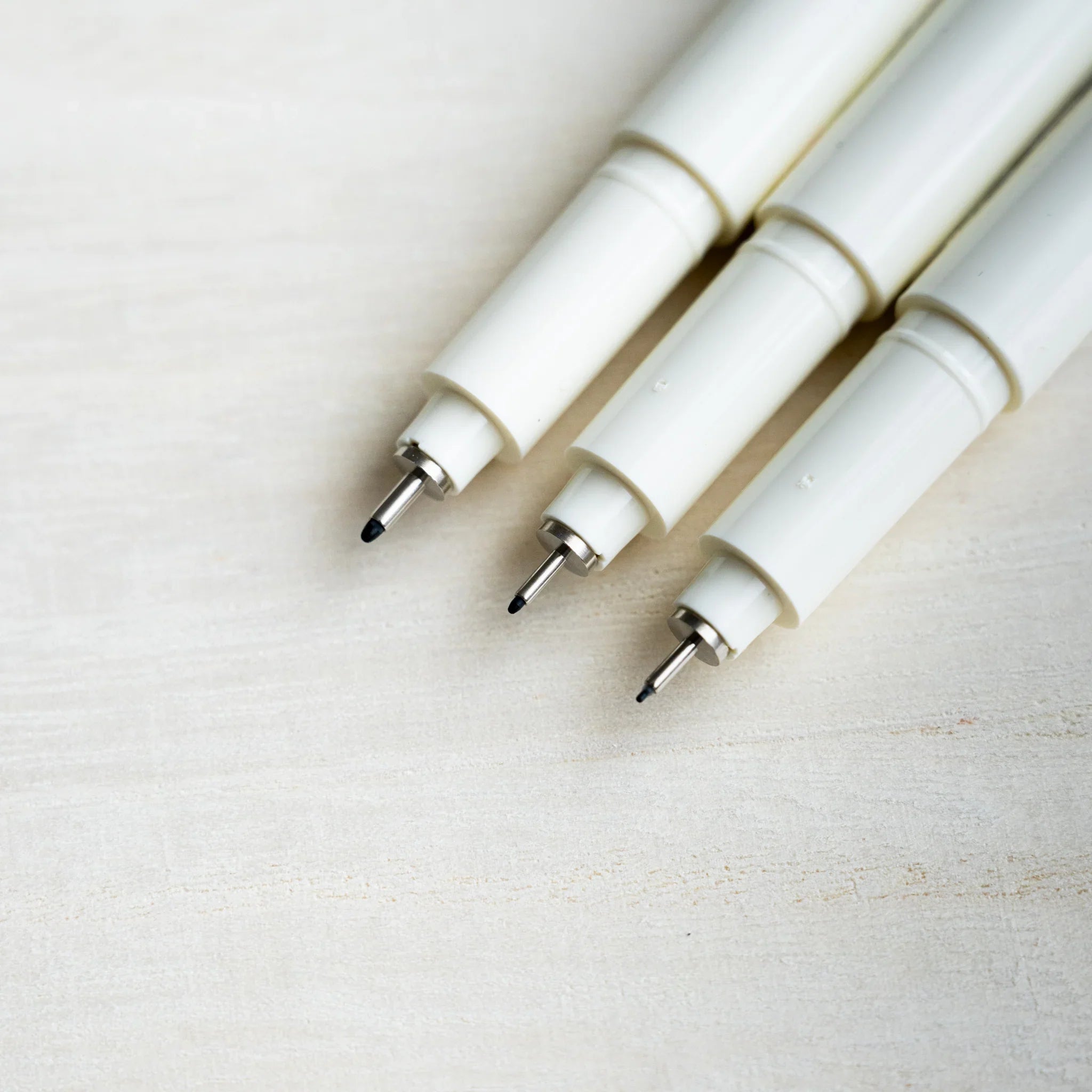 Close-up of three Midori MD Drawing Pen fine black tips with metal collars, ivory plastic barrels, on pale wood