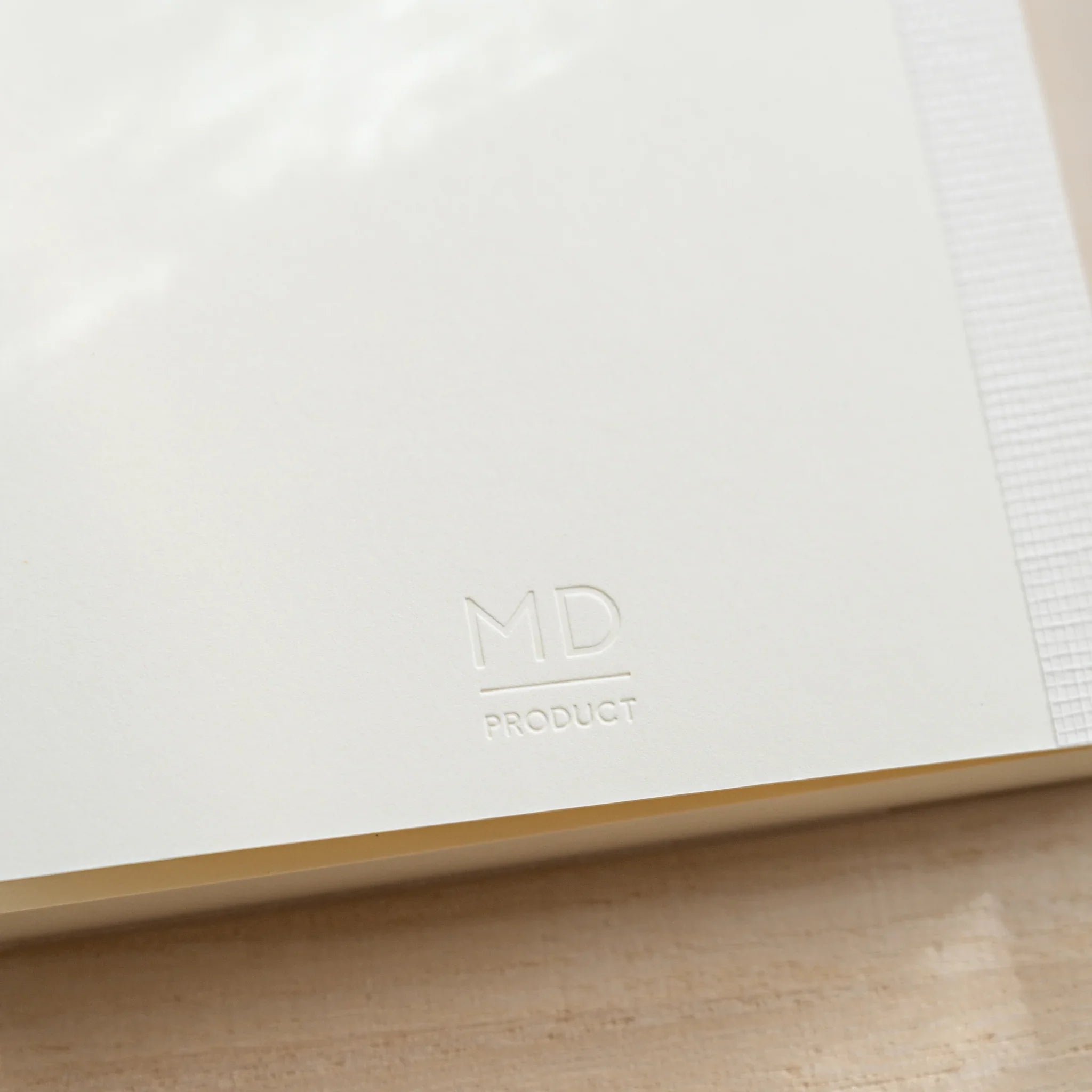 Midori MD Notebook A6 Grid corner close-up, embossed MD Product logo on white glassine, thread-stitched spine at right edge