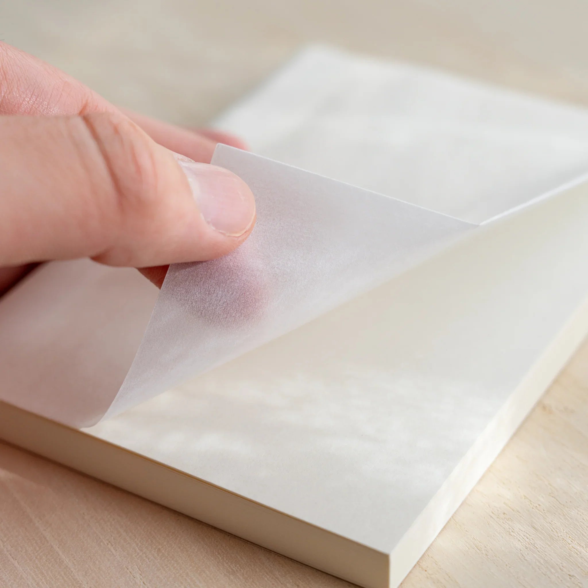 Hand lifting glassine of Midori MD Notebook A6 Grid, teal grid illustration visible through cover, ivory pages underneath