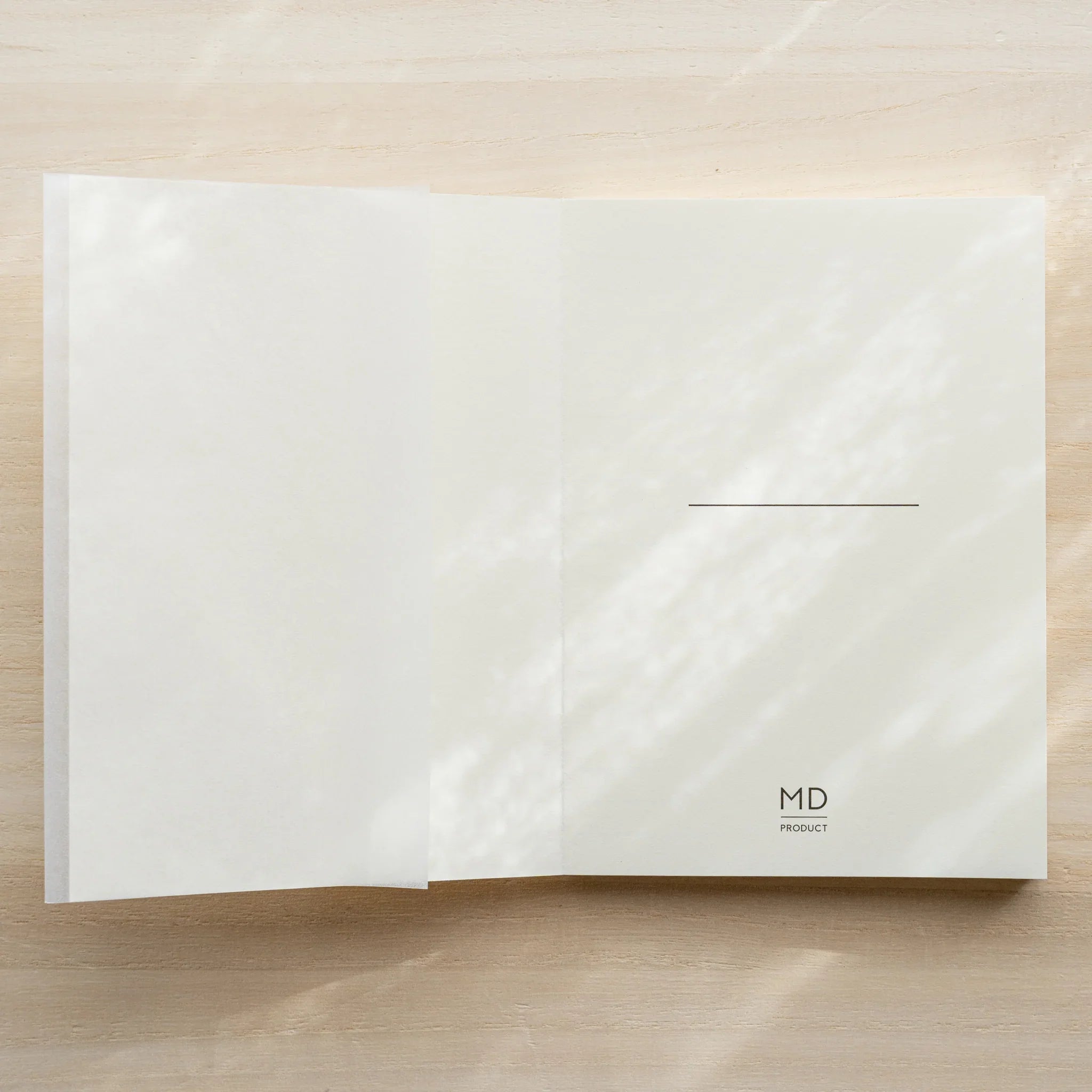 Midori MD Notebook A6 Lined with glassine cover unfolded flat beside closed notebook, MD Product logo, on wooden surface