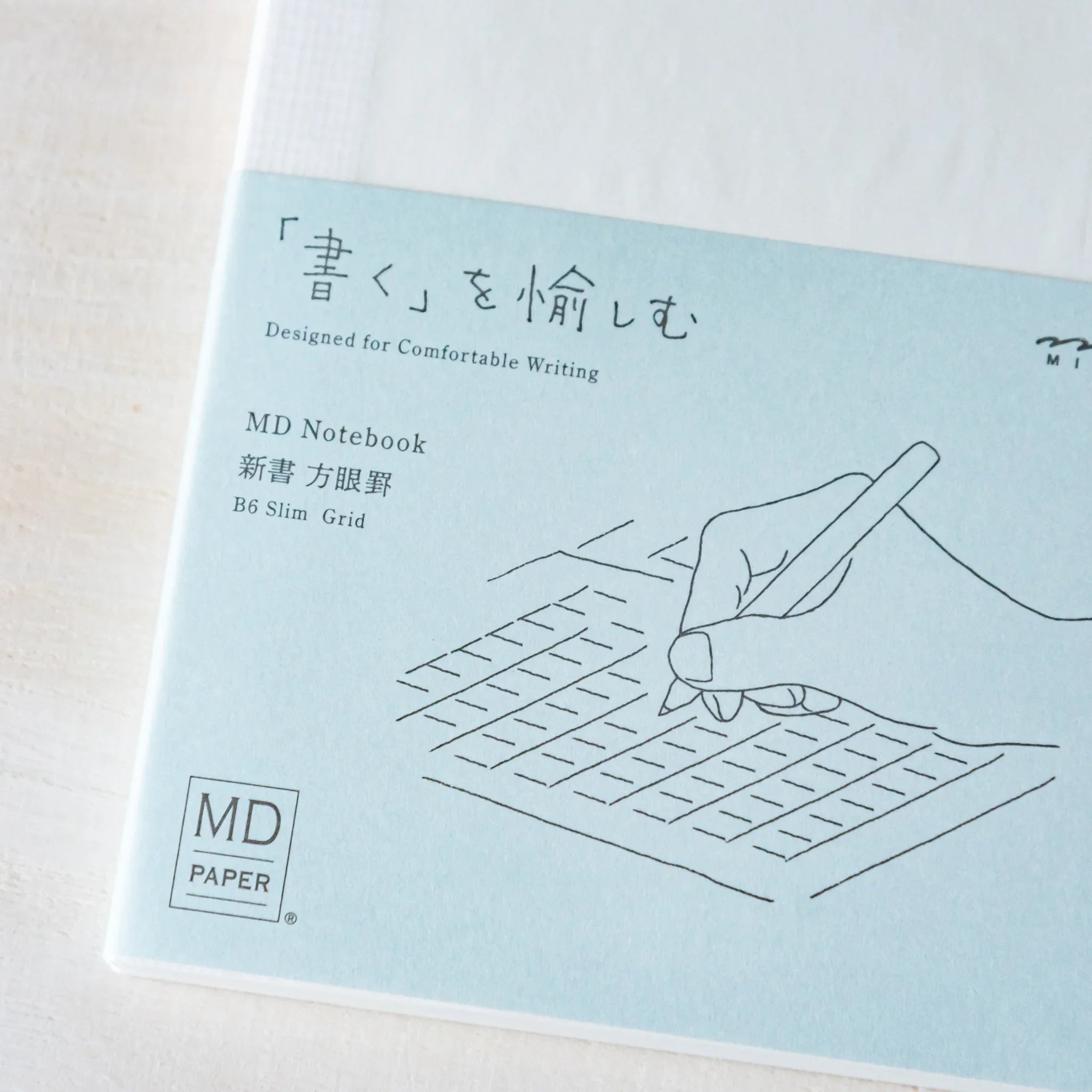 Japanese Notebook front band view of a Midori MD B6 notebook with light blue paper wrap and writing illustration.