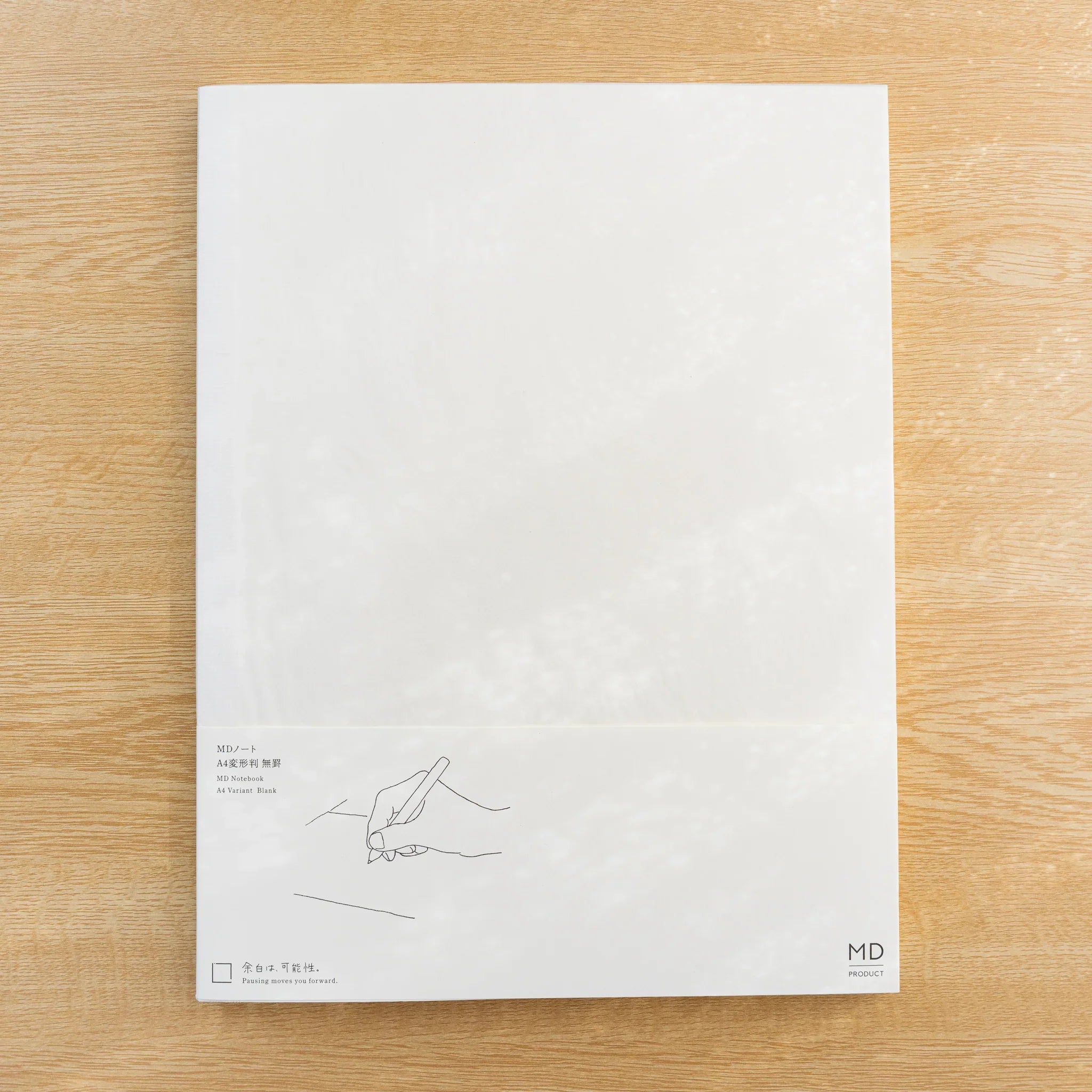 Midori MD Notebook Blank A4 Variant, cream paper cover with belly band showing a line drawing of a writing hand, on wood desk