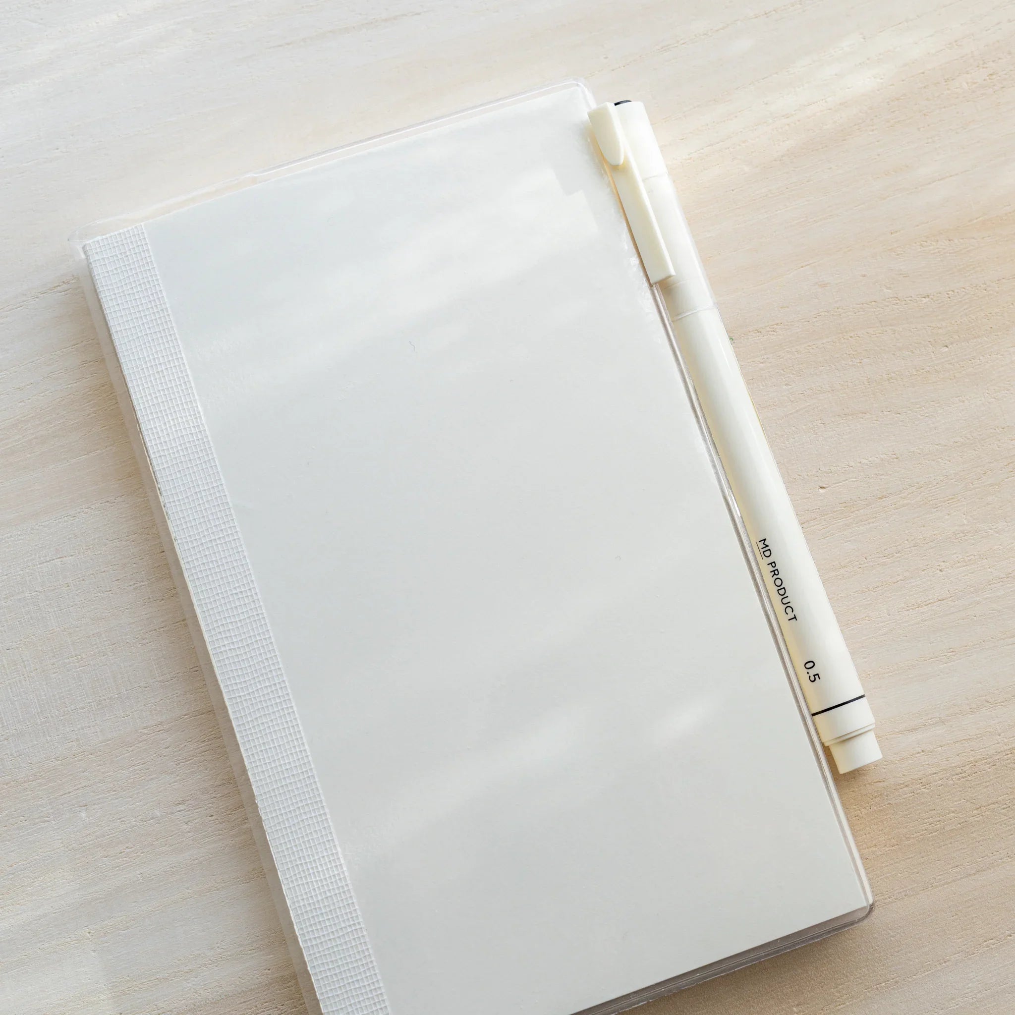 Midori MD Notebook B6 Slim in clear PVC cover with white MD PRODUCT 0.5mm pen resting diagonally across it on wooden surface