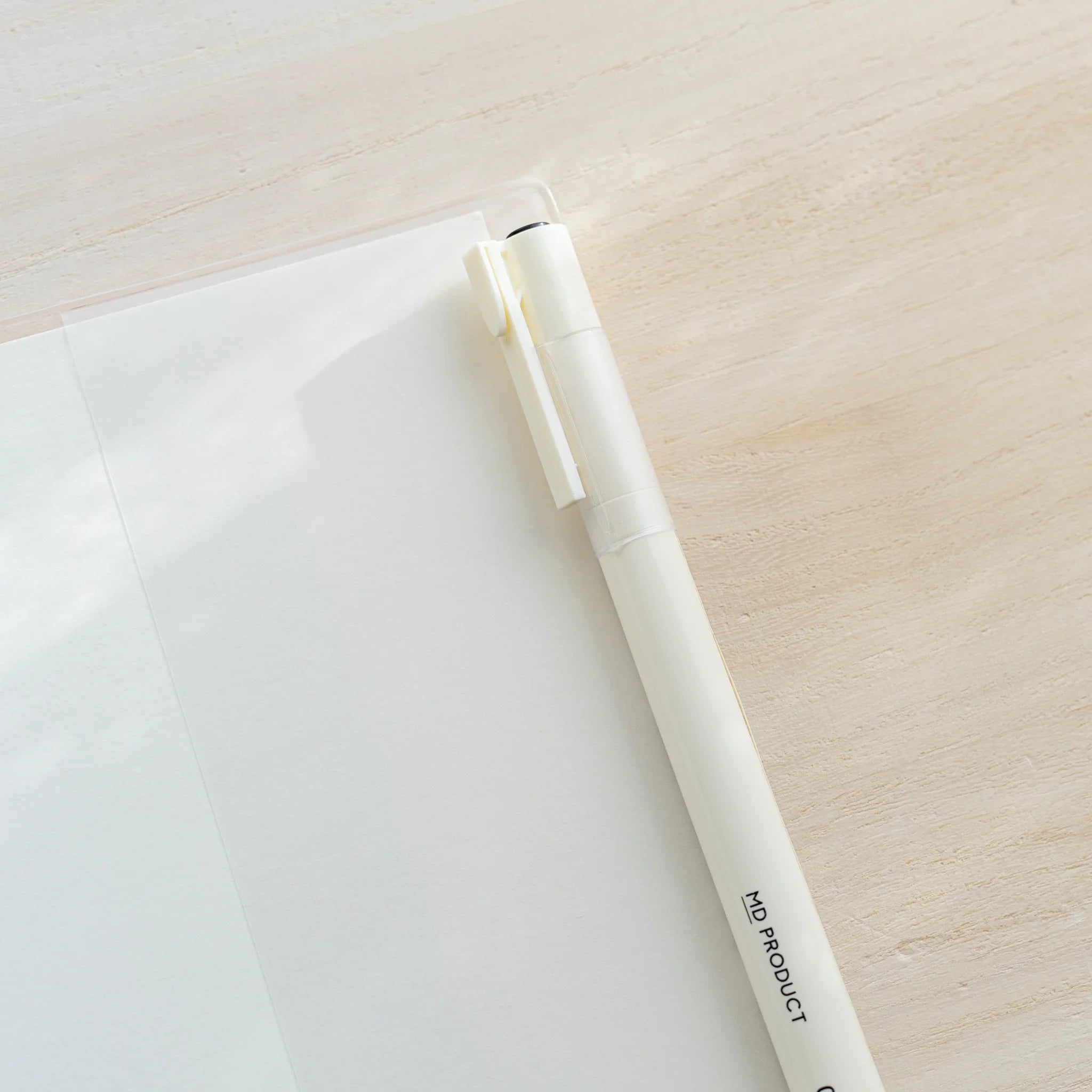 Close-up of white MD PRODUCT pen resting across corner of Midori MD Notebook Cover clear PVC on wooden surface