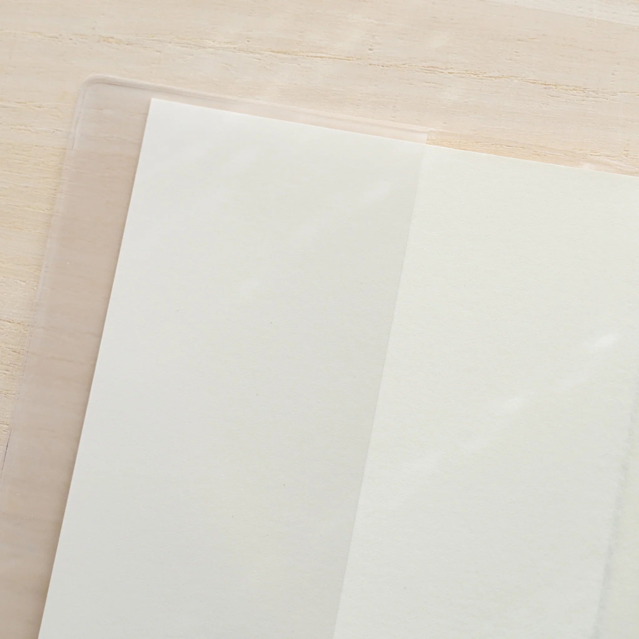 Close-up of MD Notebook Cover A5 top-left corner showing clear PVC edge overlapping white notebook cover on wooden surface