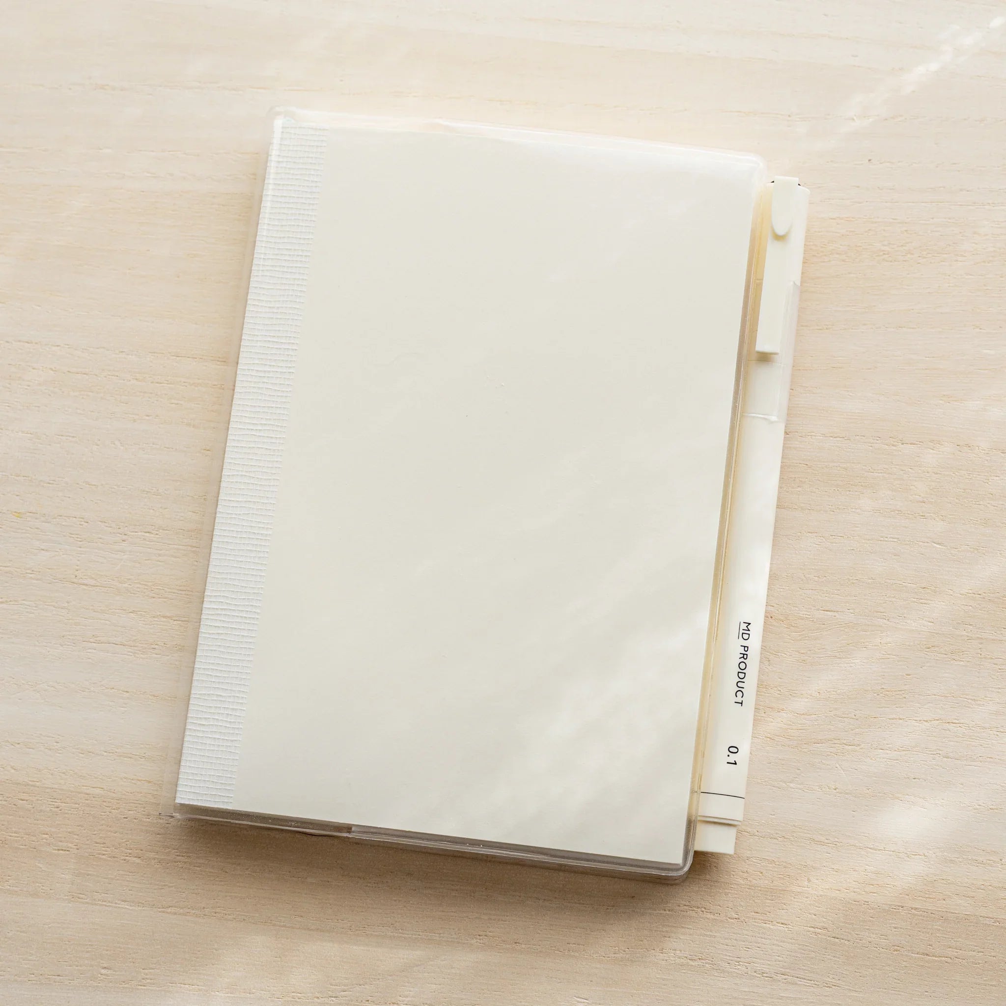 Midori MD Notebook A5 in clear PVC cover, closed front view on wooden surface, white pen in right-side pen holder