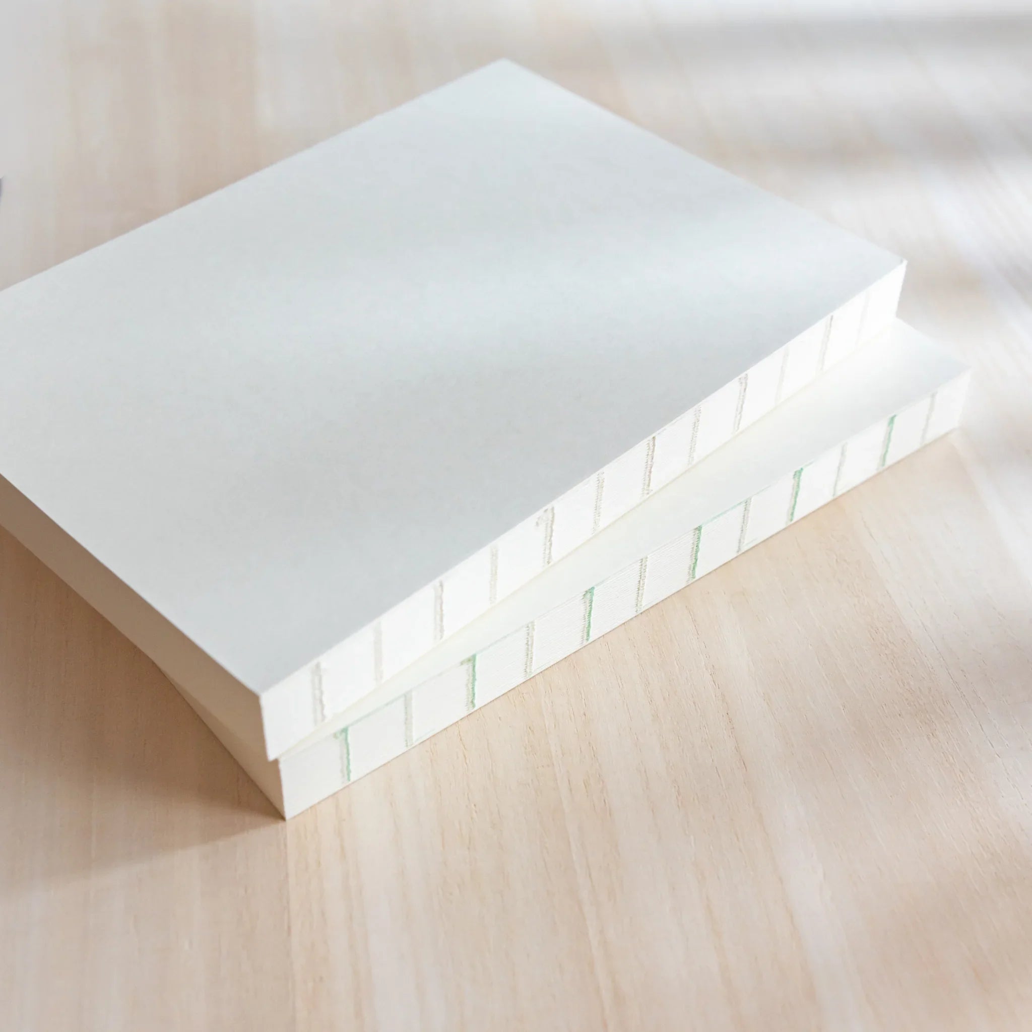 Angled view of two stacked Midori Japanese journals with plain white covers and grid spine on light wood.