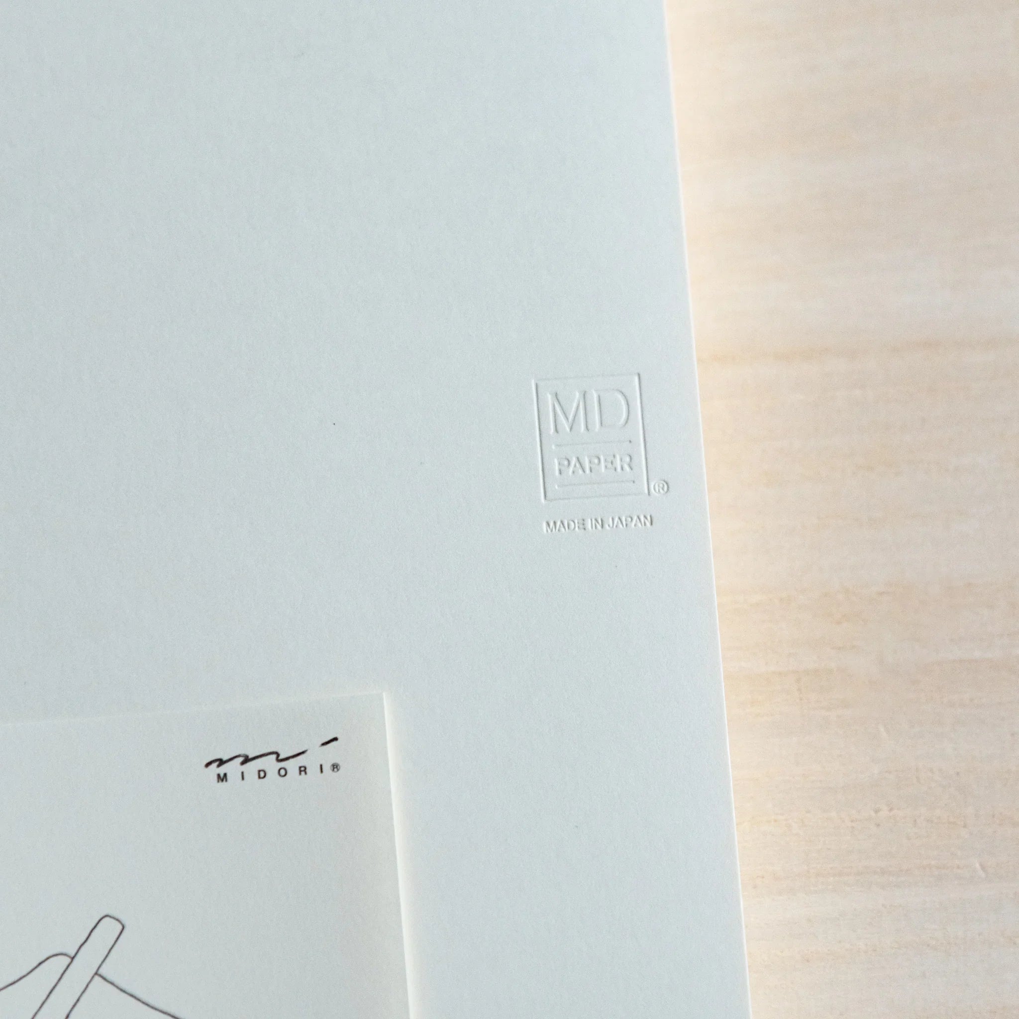 Close-up of Midori Japanese journal with white cover, embossed MD logo and small illustration card on wood.