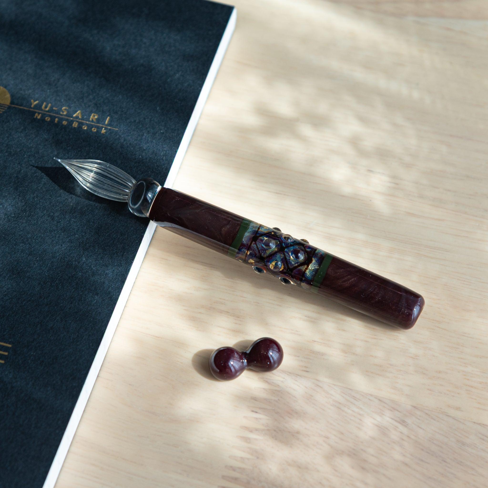 A glass dip pen handmade in Japan with a maroon body and decorative band, resting on a wooden desk near a notebook.