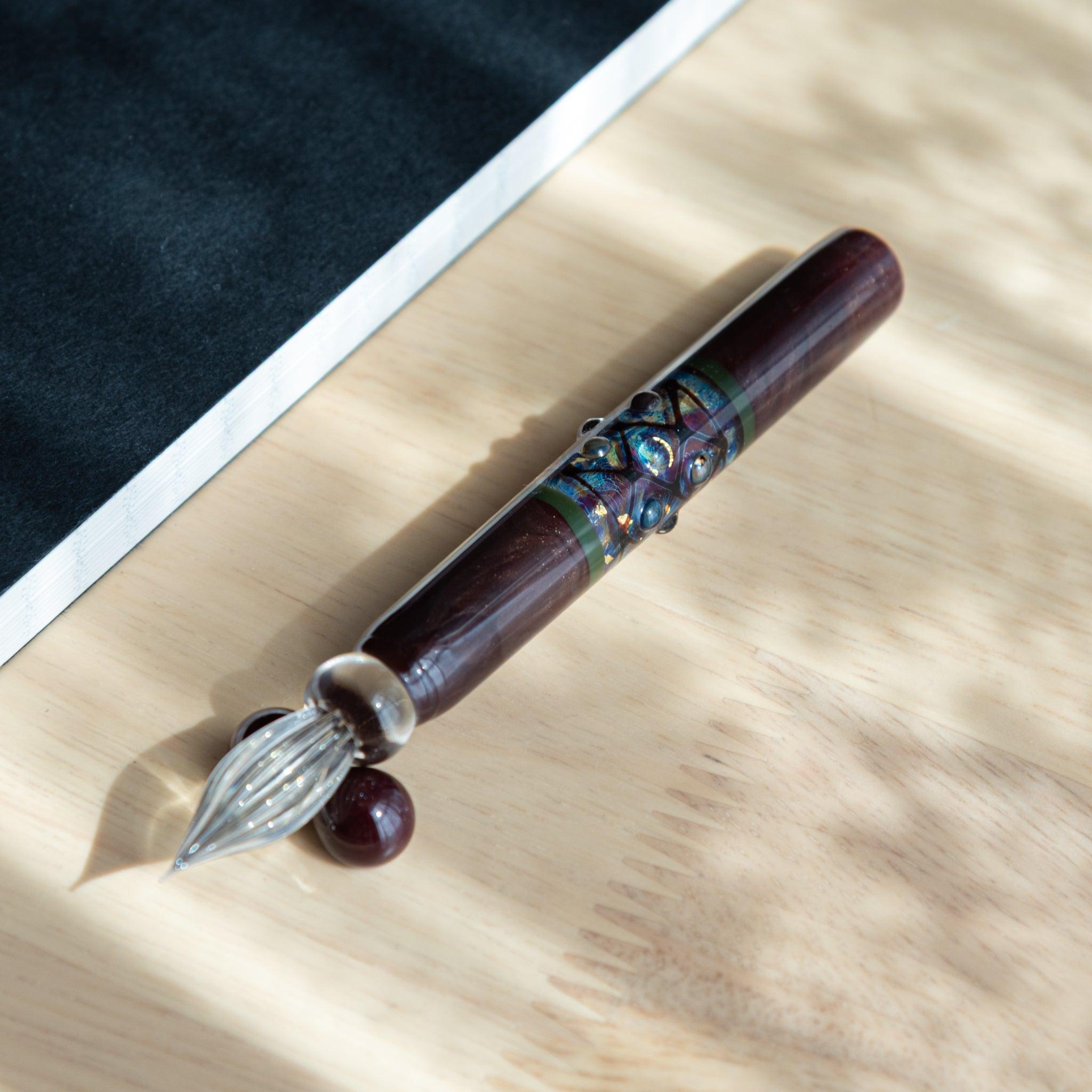 A Japanese glass dip pen with a glossy maroon body and intricate detailing, placed diagonally on a wooden surface.