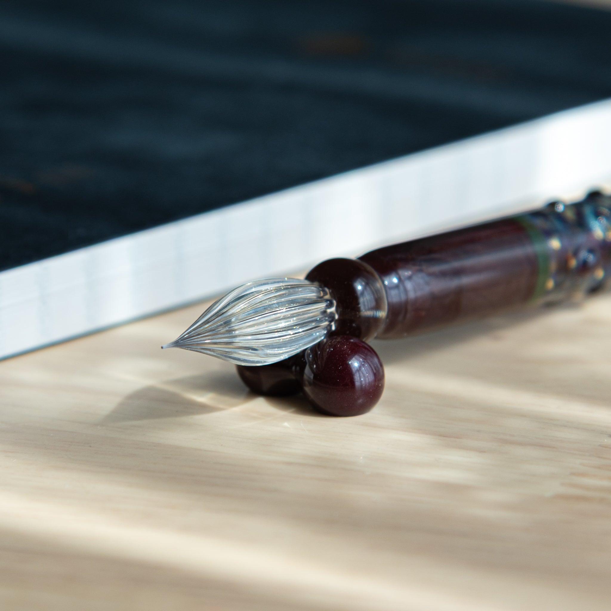 Close-up of a Japanese glass dip pen with a maroon body and clear twisted nib, resting on a wooden desk near a notebook.
